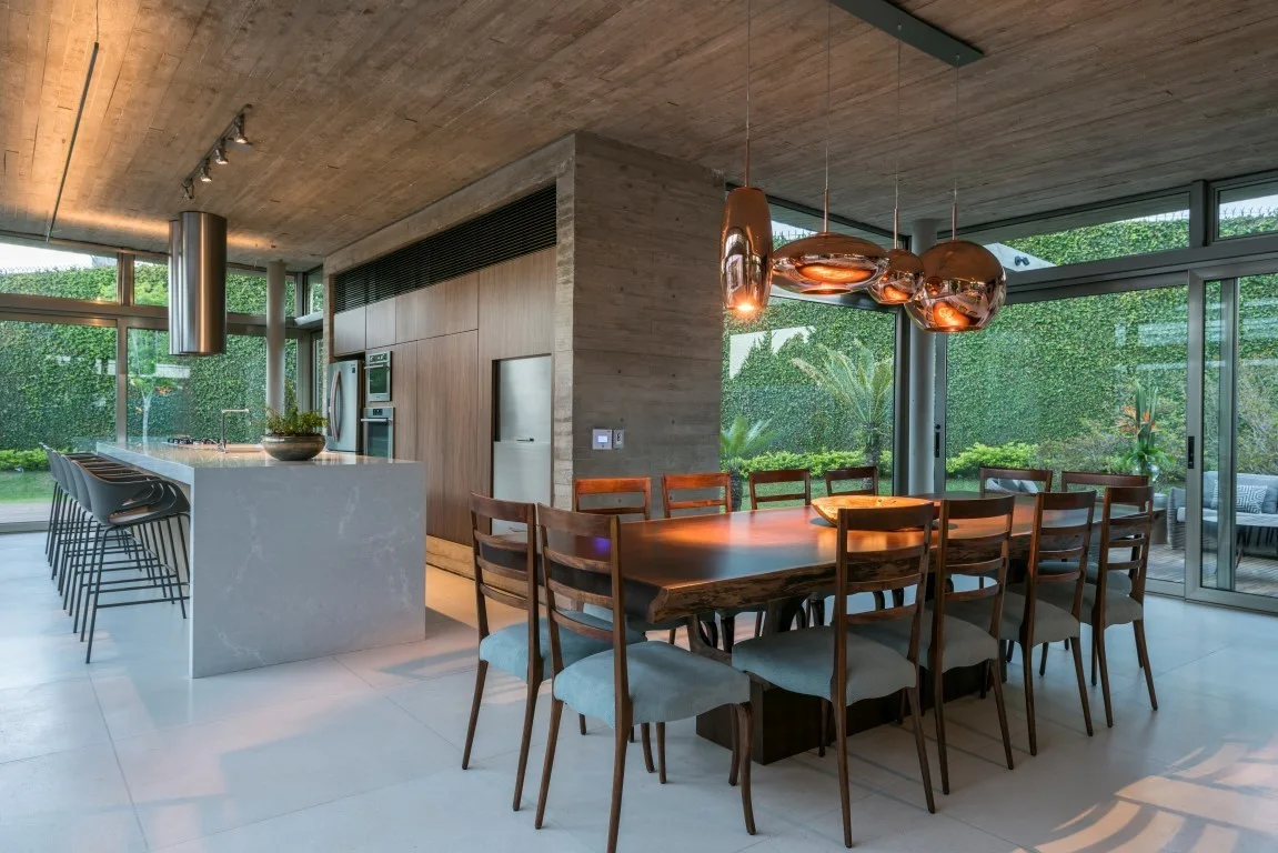 A modern open-plan dining room featuring a live-edge wood table, copper pendant lights, a white kitchen island, and a textured concrete ceiling.
