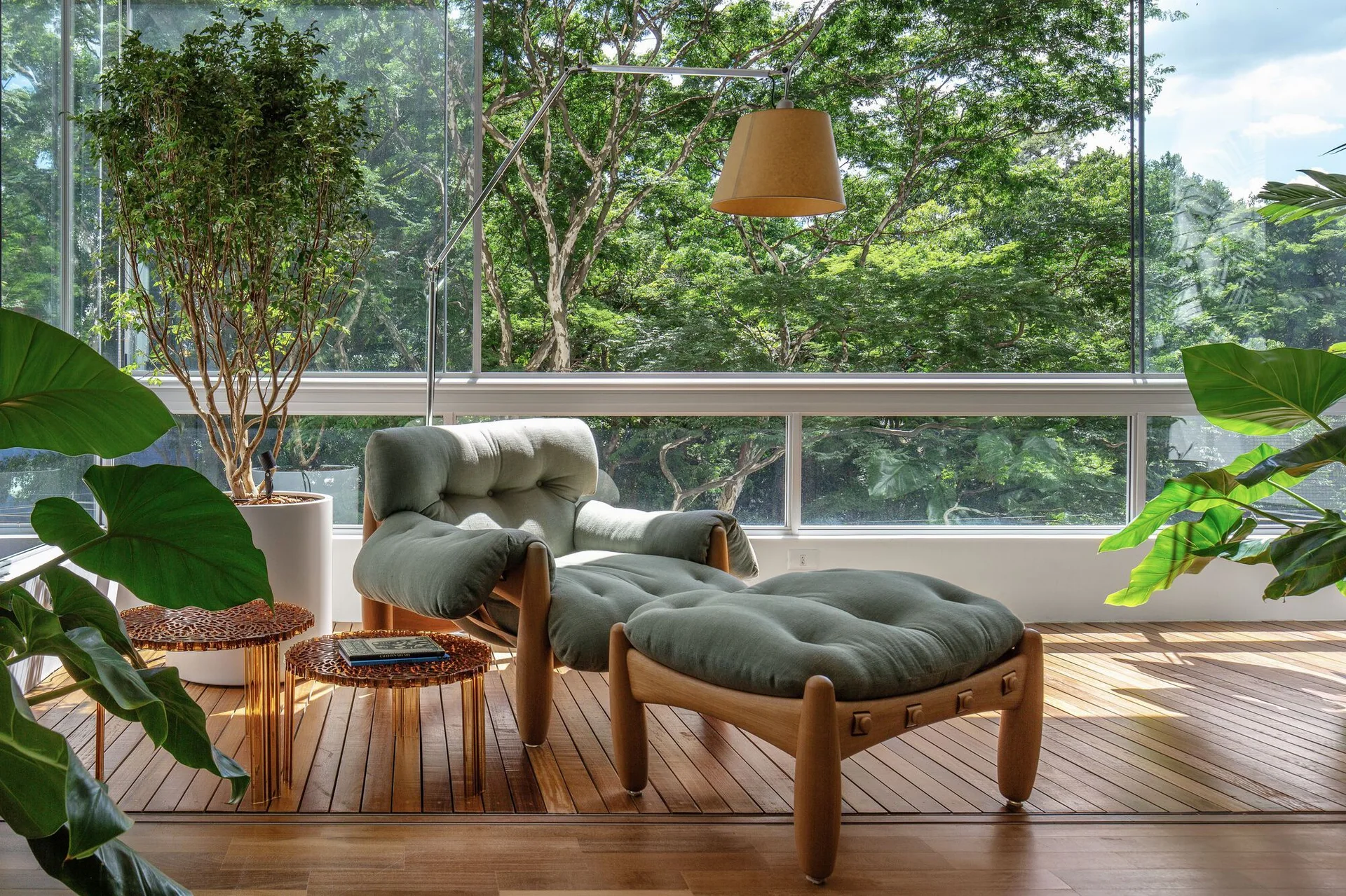 A tufted sage green lounge chair and matching ottoman sit on a wood plank floor in front of a floor-to-ceiling glass wall showing green trees.