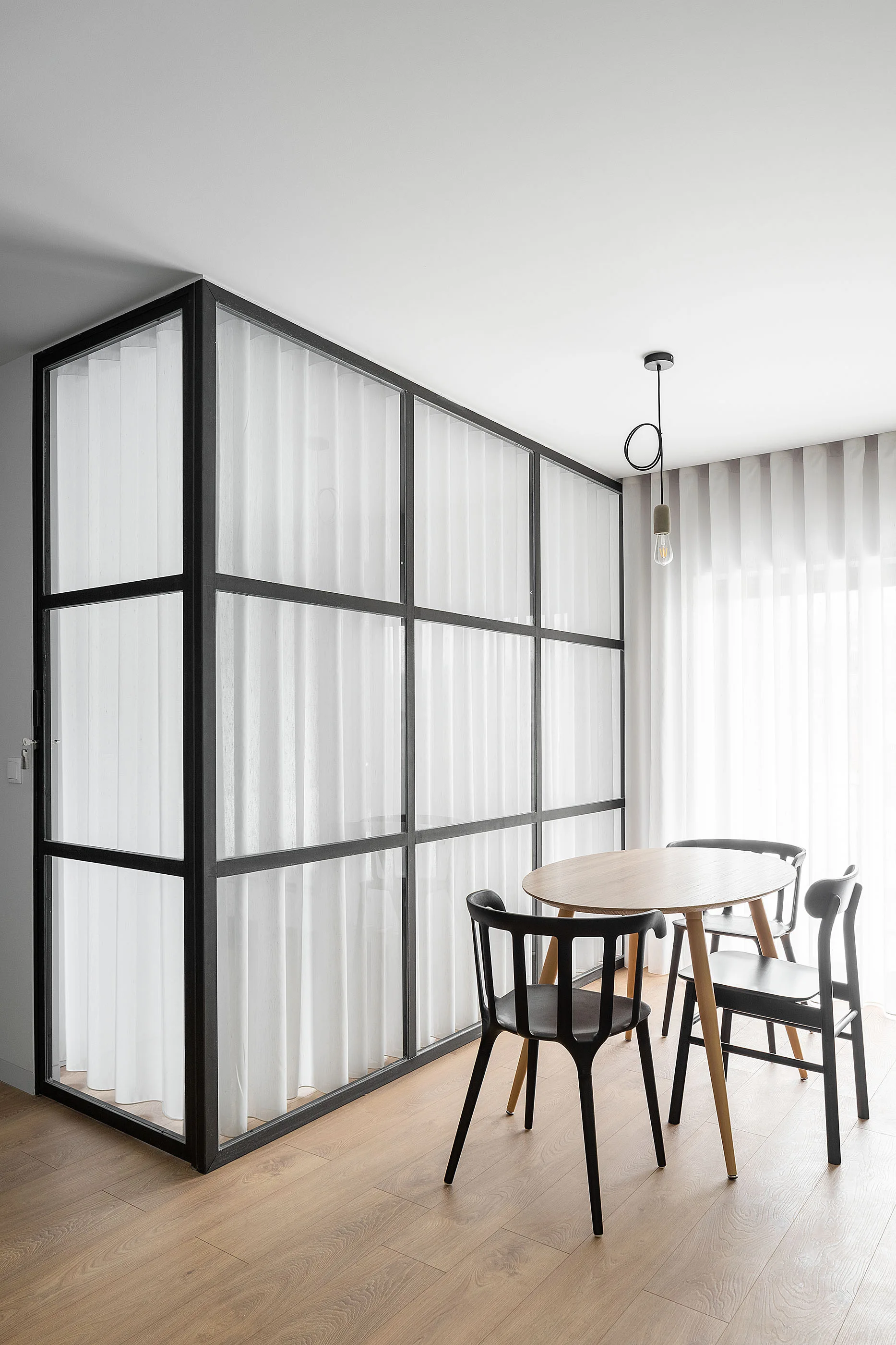 A minimalist dining area featuring a round wooden table, black chairs, and a black steel grid room partition lined with white sheer curtains.