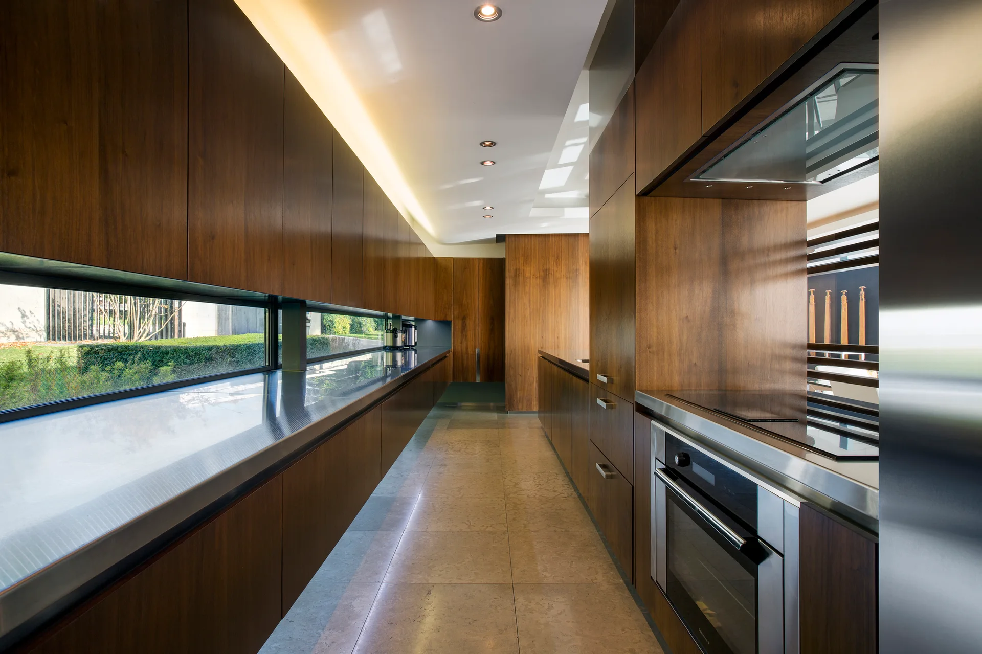 A modern galley kitchen features floor-to-ceiling wood cabinetry, stainless steel countertops, a long horizontal ribbon window, and tile flooring.