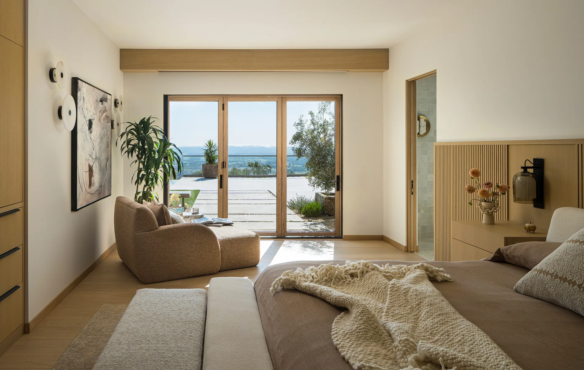A neutral bedroom features a bed with a chunky knit throw, a slatted wood headboard, a textured lounge chair, and glass doors opening to a scenic terrace.