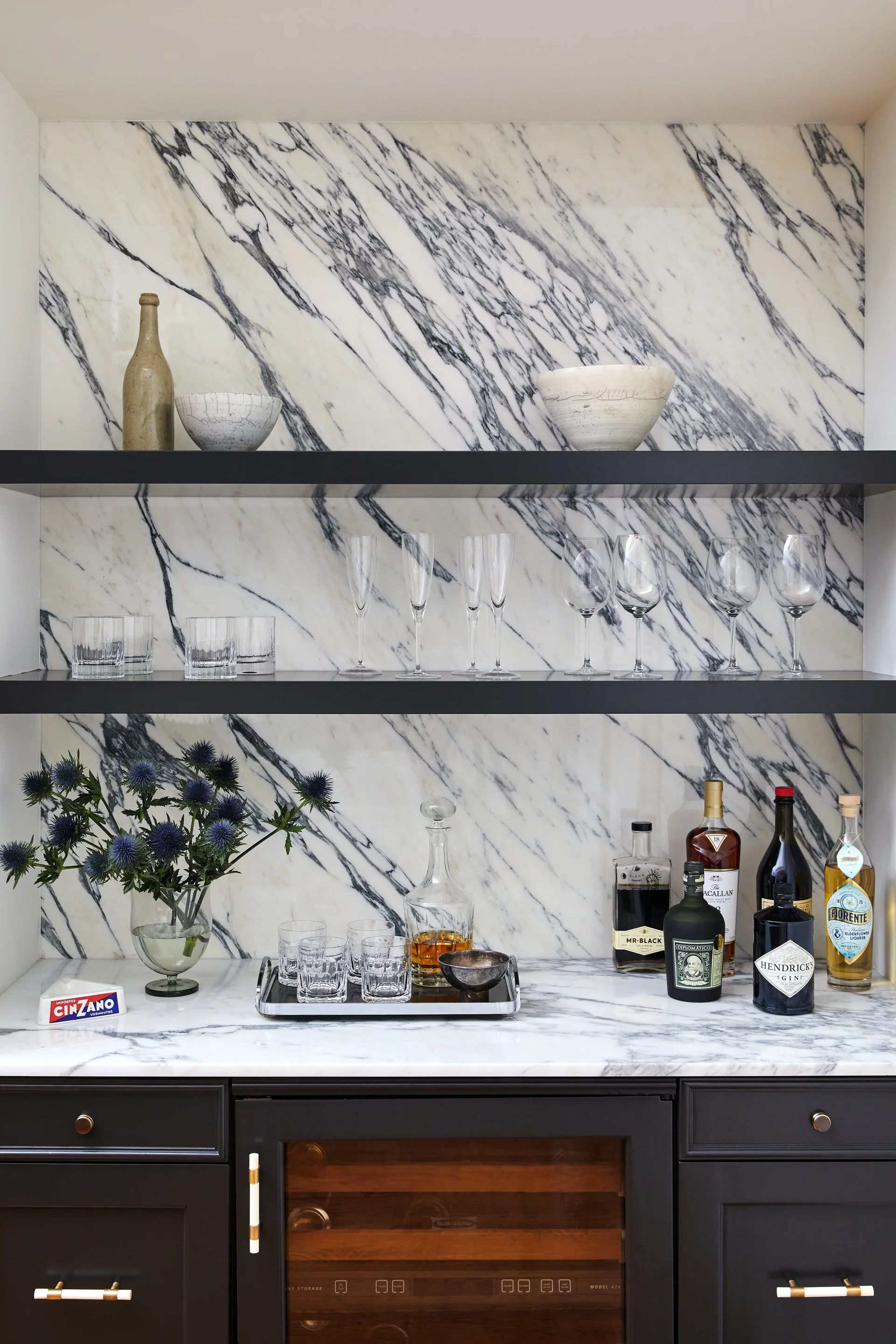 A modern home bar features a white marble backsplash with grey veining, matte black cabinets, and two black floating shelves holding glassware.