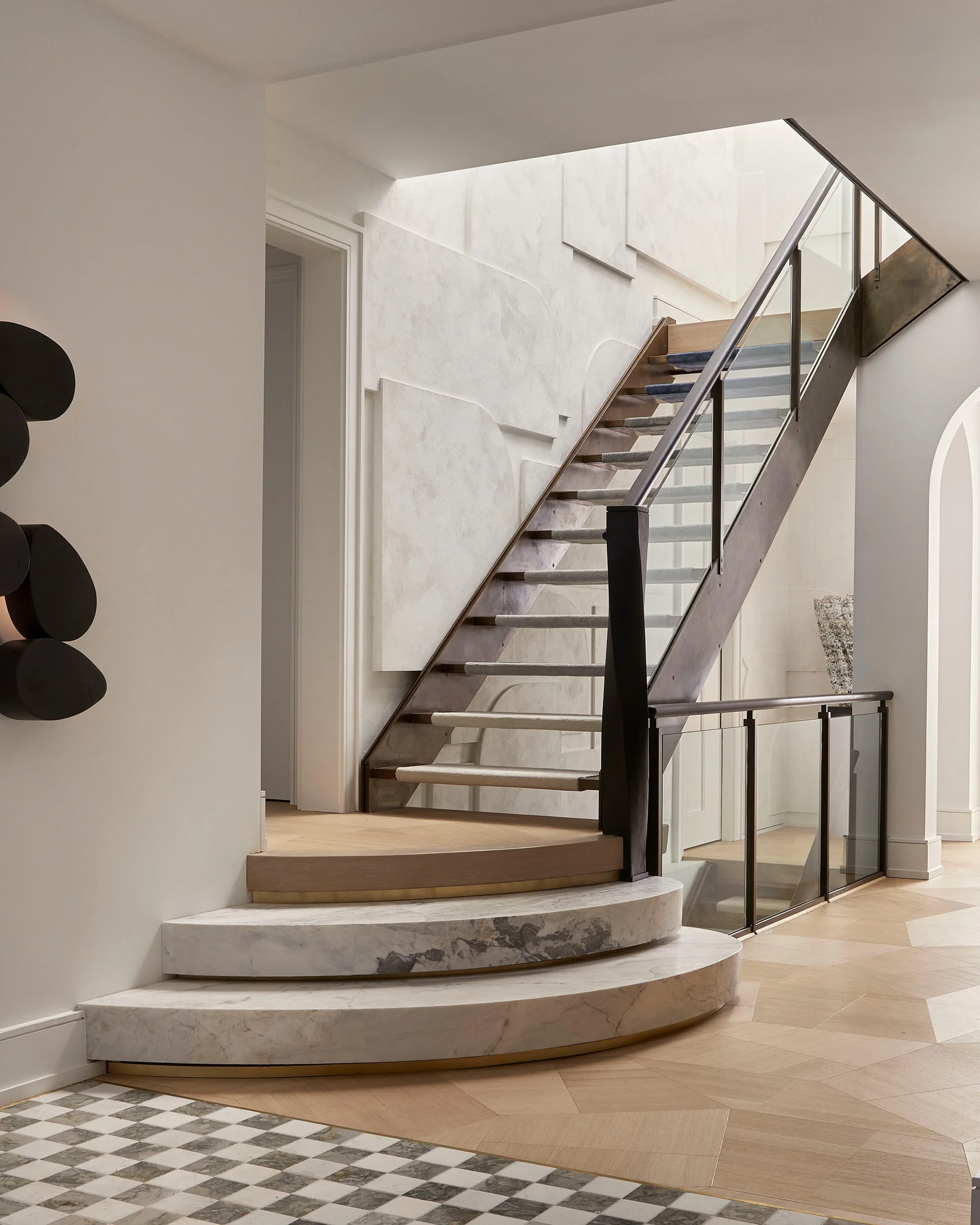 A modern staircase in a foyer features curved marble base steps, wood treads, a glass railing, and textured white geometric wall paneling.
