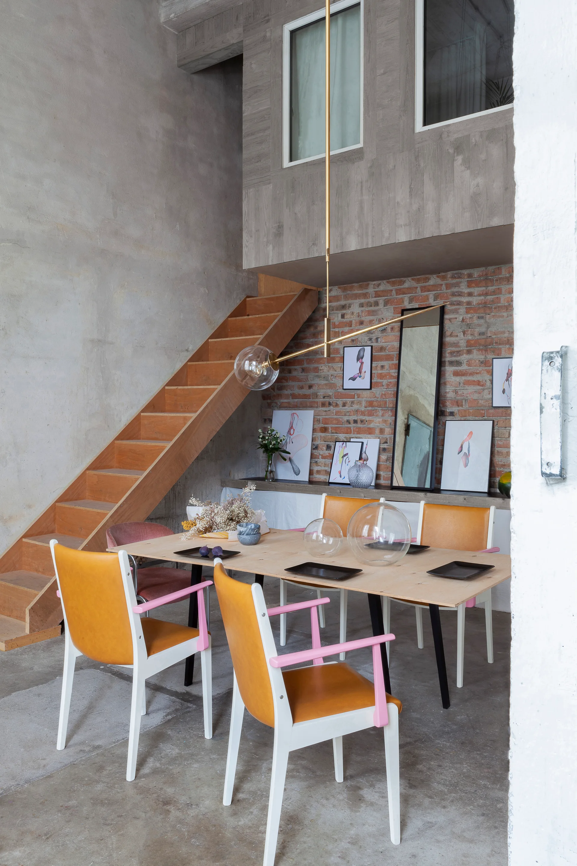 A modern loft dining room features a light wood table surrounded by yellow and pink chairs, an exposed brick wall, and a wooden staircase.