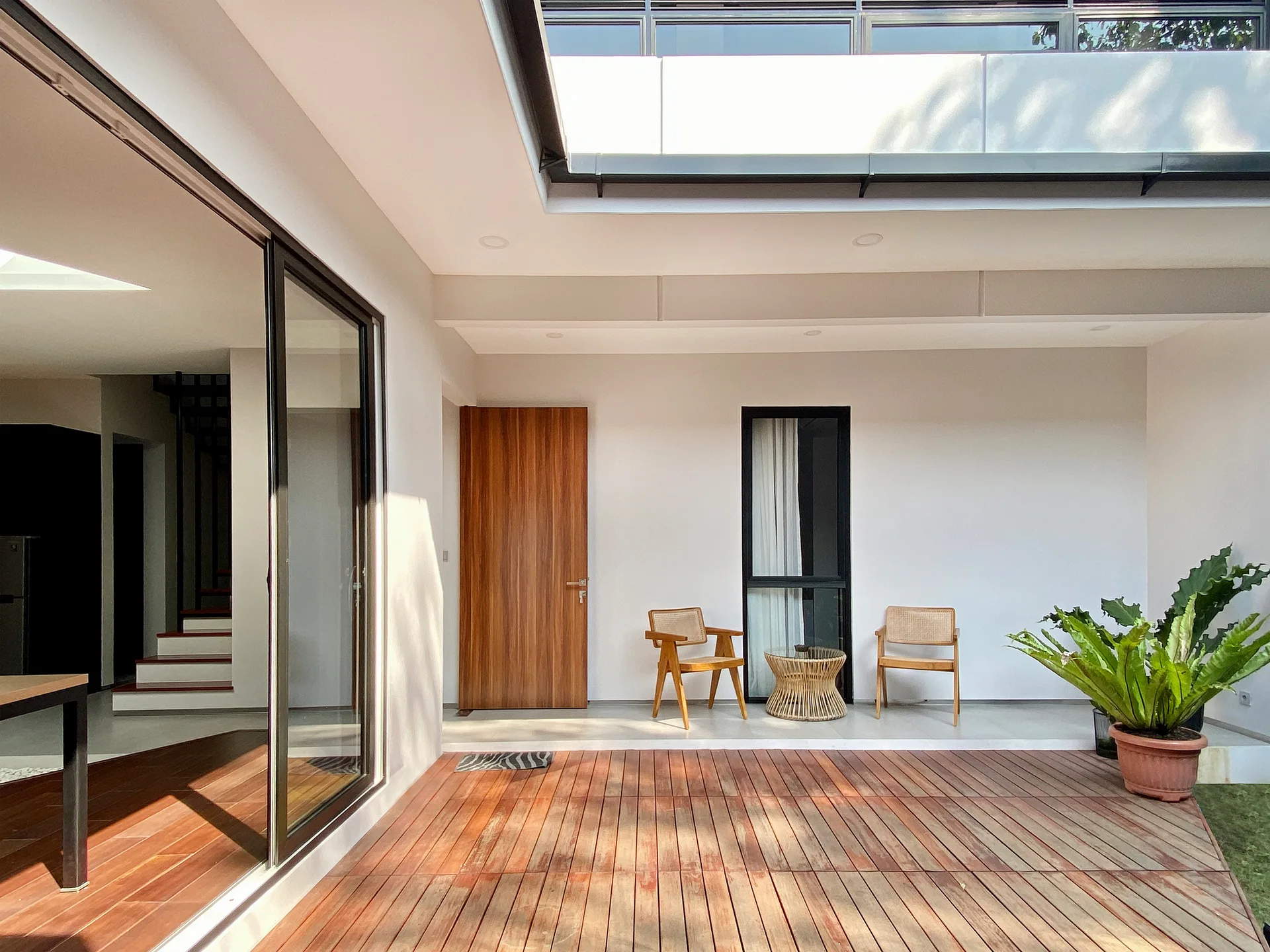 A modern outdoor patio featuring a warm wood deck, two woven chairs with a small table against a white wall, and a large sliding glass door.