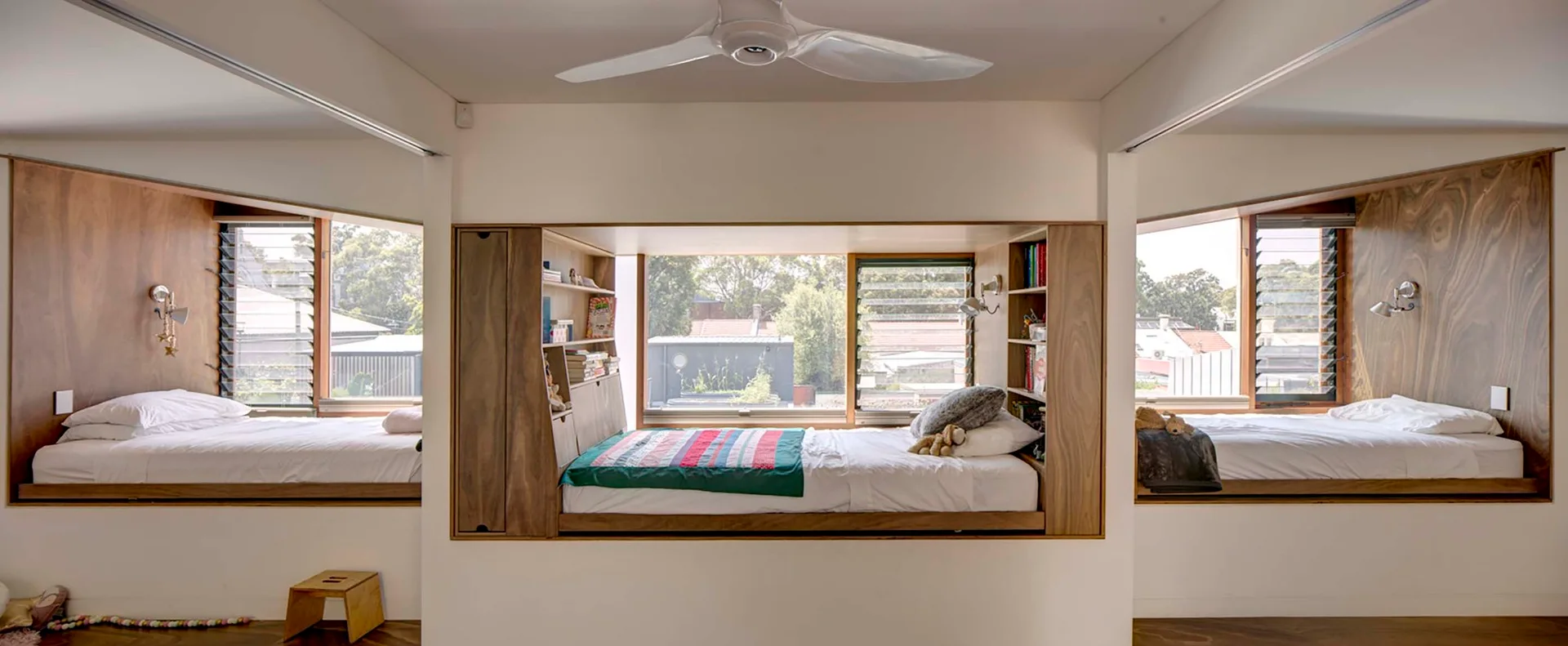 A custom wooden bed niche built into a white wall features integrated bookshelves, a large window, and cozy bedding.