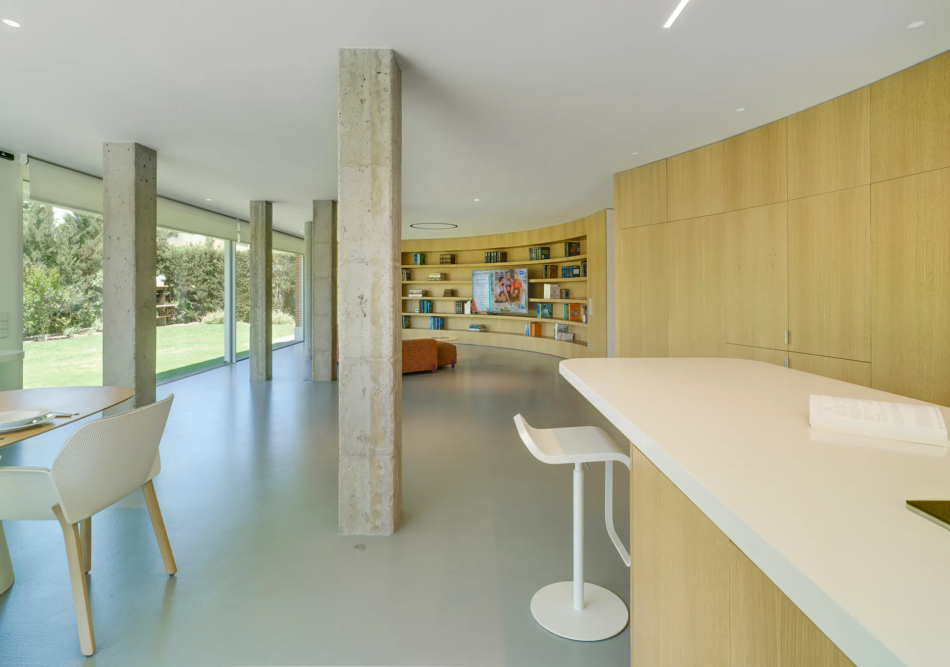 An open-concept living space features exposed concrete columns, a curved wood-paneled bookshelf wall, and a white kitchen island.