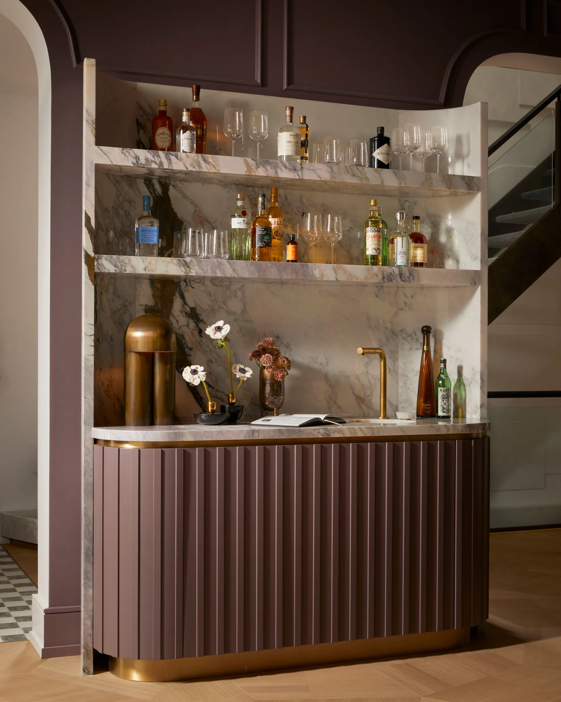 A custom home bar features a plum-colored fluted cabinet under a veined white marble niche with matching stone shelving and a brass faucet.