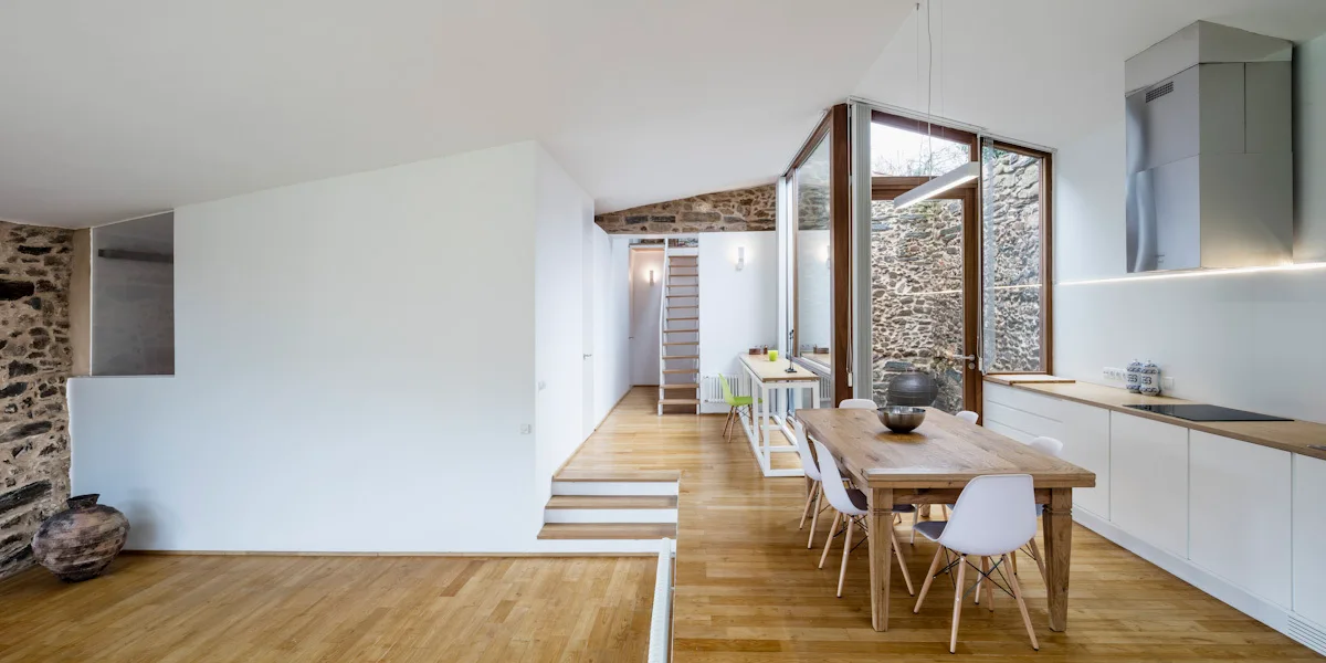 A modern open-concept kitchen and dining space with white cabinetry, a wood dining table, wood flooring, and exposed natural stone walls.