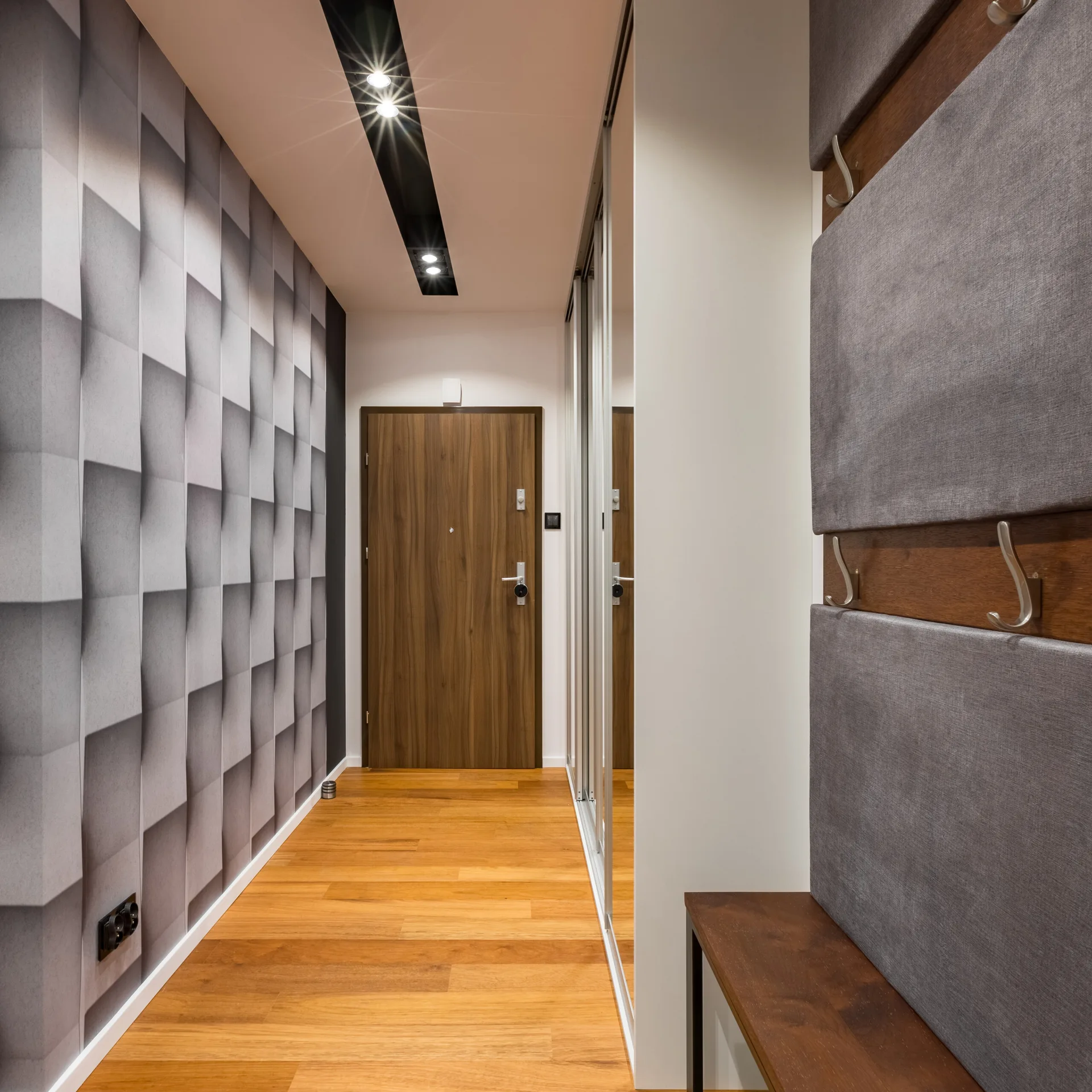 A modern entryway featuring 3D-patterned grey wallpaper, a wooden floor, and a built-in bench with grey upholstered wall panels and metal hooks.