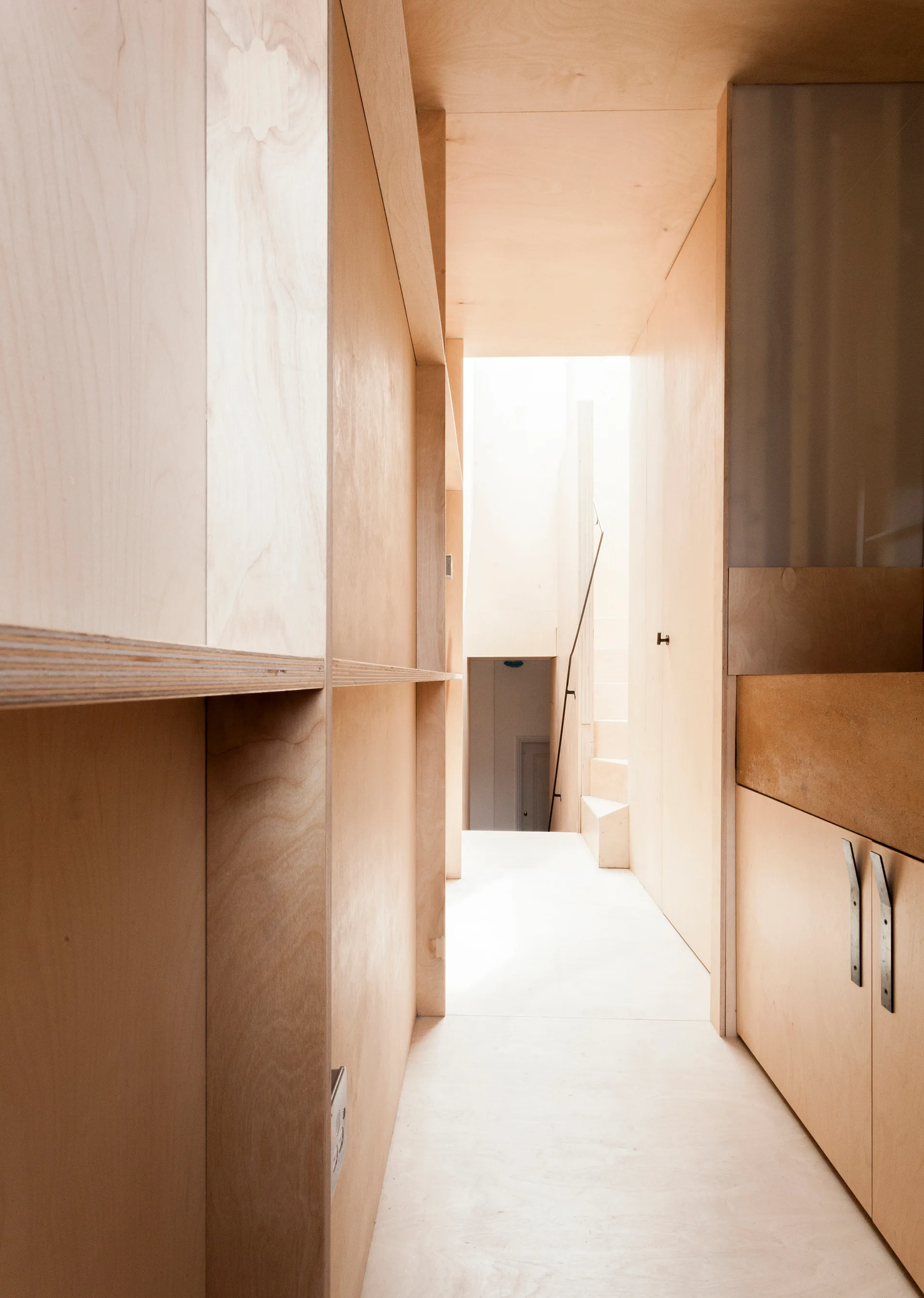 A narrow hallway features light tan plywood wall paneling, built-in storage cabinets, and a staircase visible in the brightly lit background.