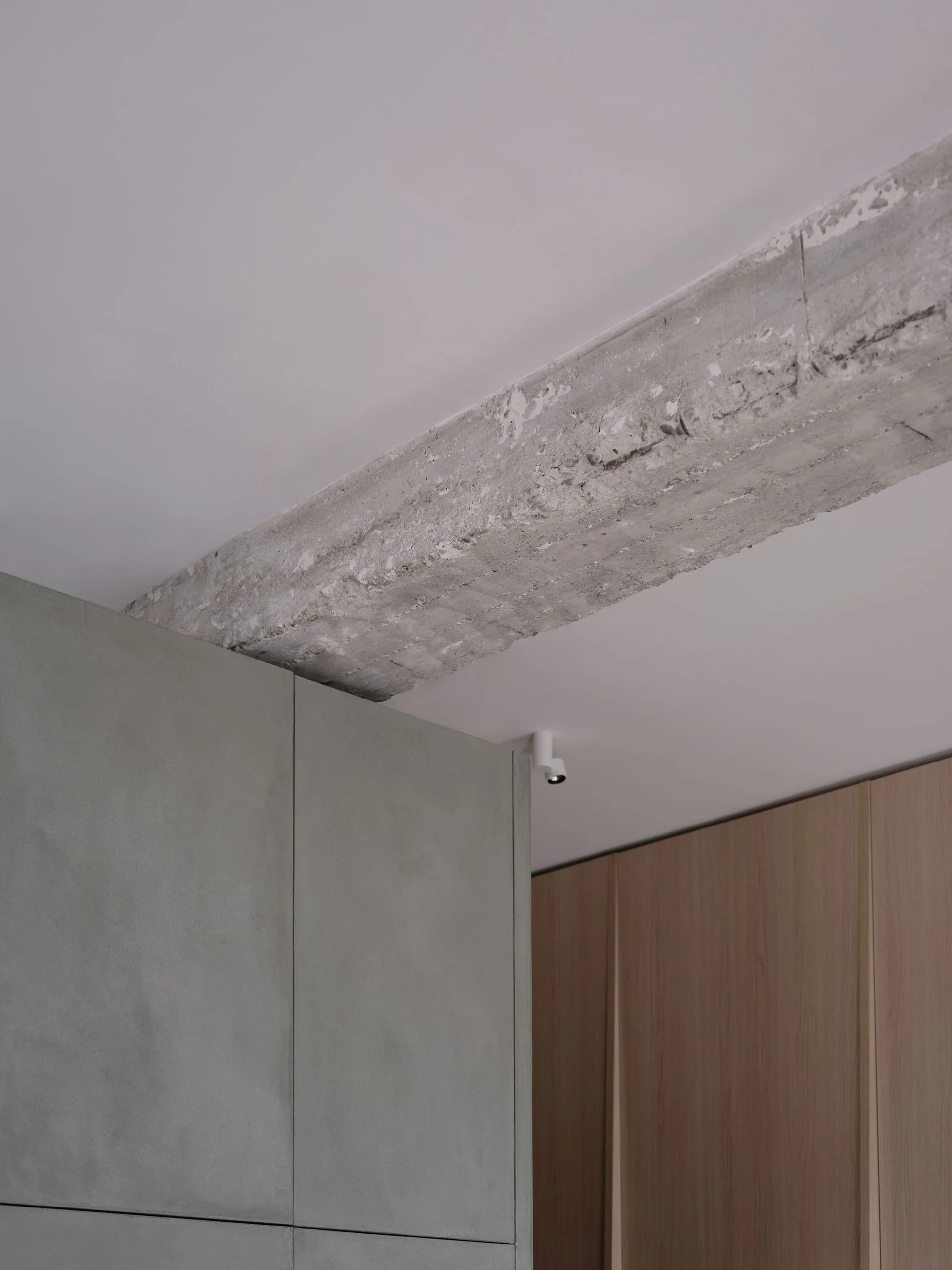 A close-up of a raw concrete beam across a white ceiling above matte sage green and light wood flat-panel cabinets.
