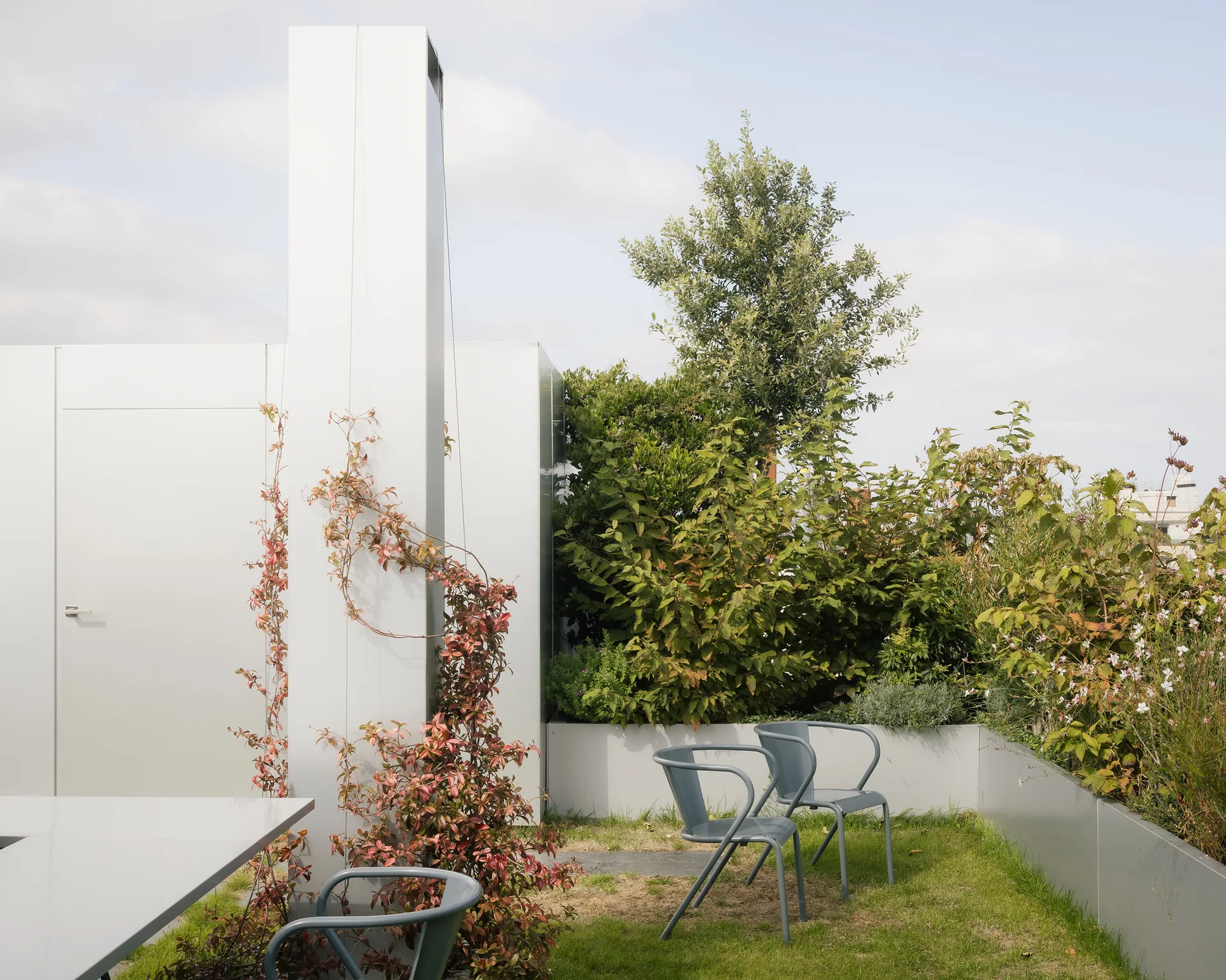 Two gray metal chairs sit on a small grass lawn on a modern rooftop terrace surrounded by built-in gray planters and metal walls.