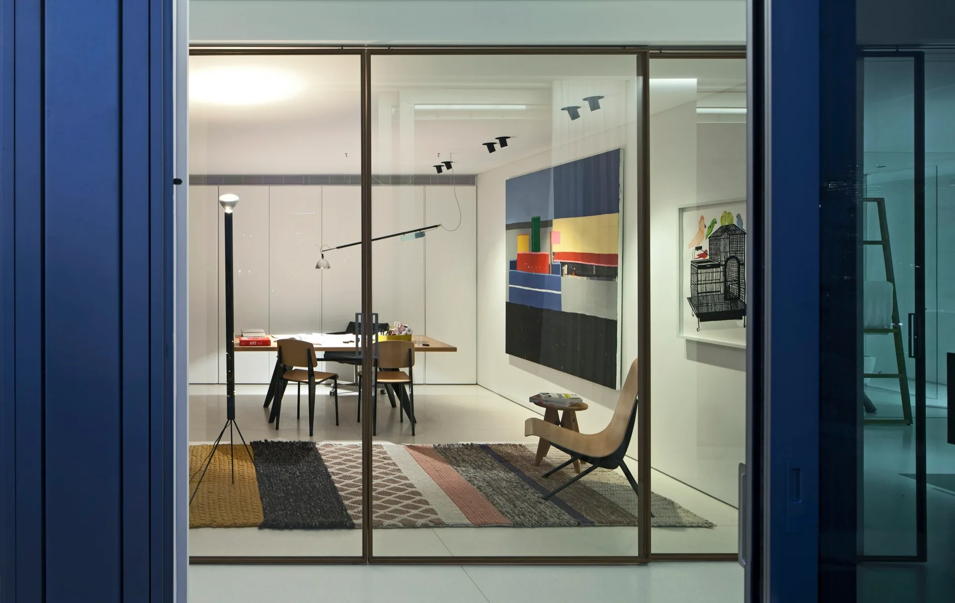A modern home office with a wooden desk and a curved lounge chair is visible through large sliding glass doors, featuring a colorful textured rug.