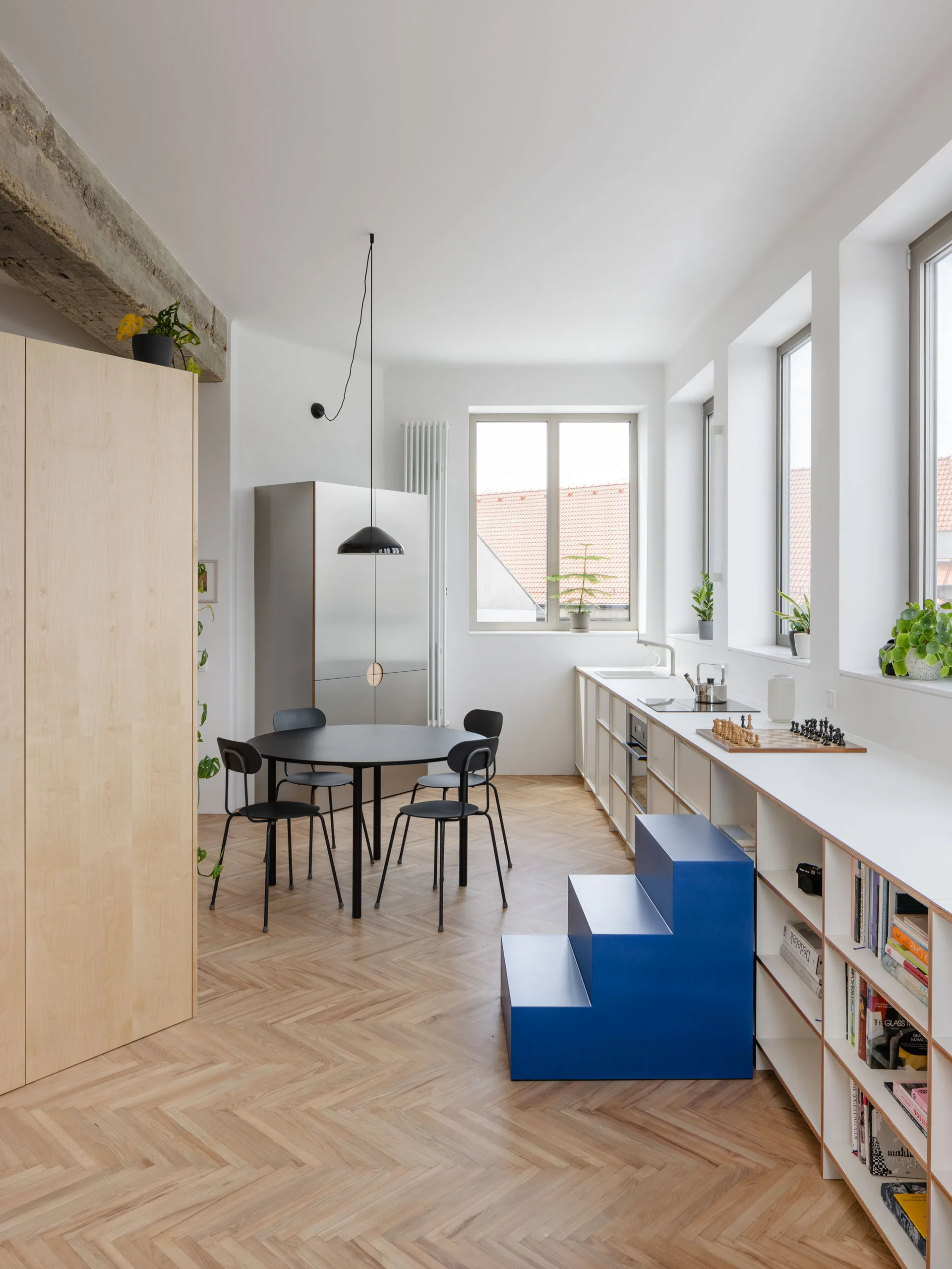 A minimalist kitchen features a light wood herringbone floor, a black dining set with a pendant light, and a bright blue stepped stool.