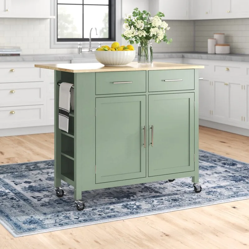 A sage green mobile kitchen island with a light wood top rests on a blue patterned rug in front of white cabinets.