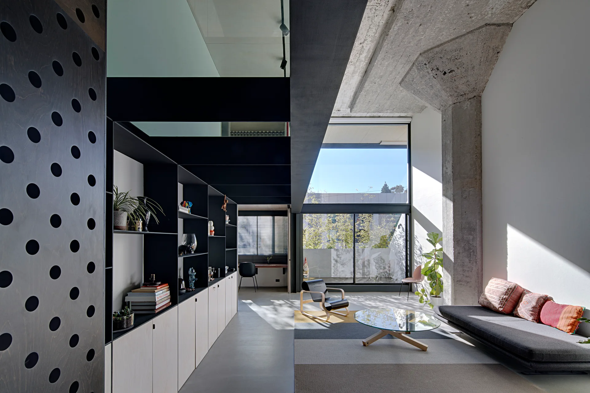 A modern living room features an exposed concrete column, a low-profile gray sofa, a glass coffee table, and large black metal shelving along the wall.