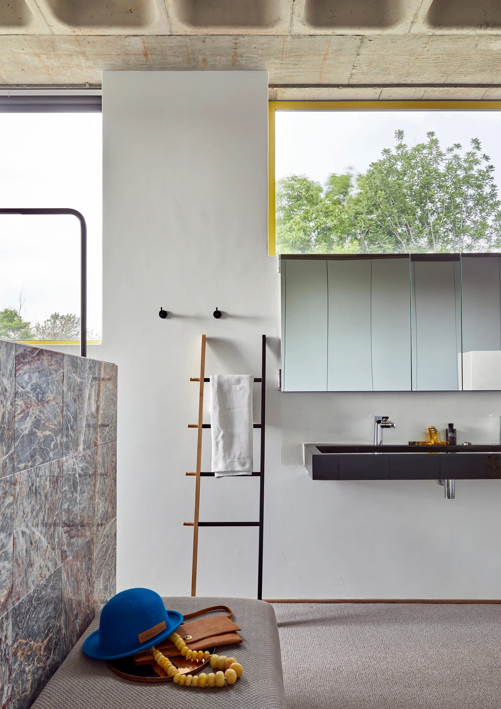 A modern bathroom features a floating black vanity sink, a leaning towel ladder, a stone partition wall, and an exposed concrete waffle ceiling.