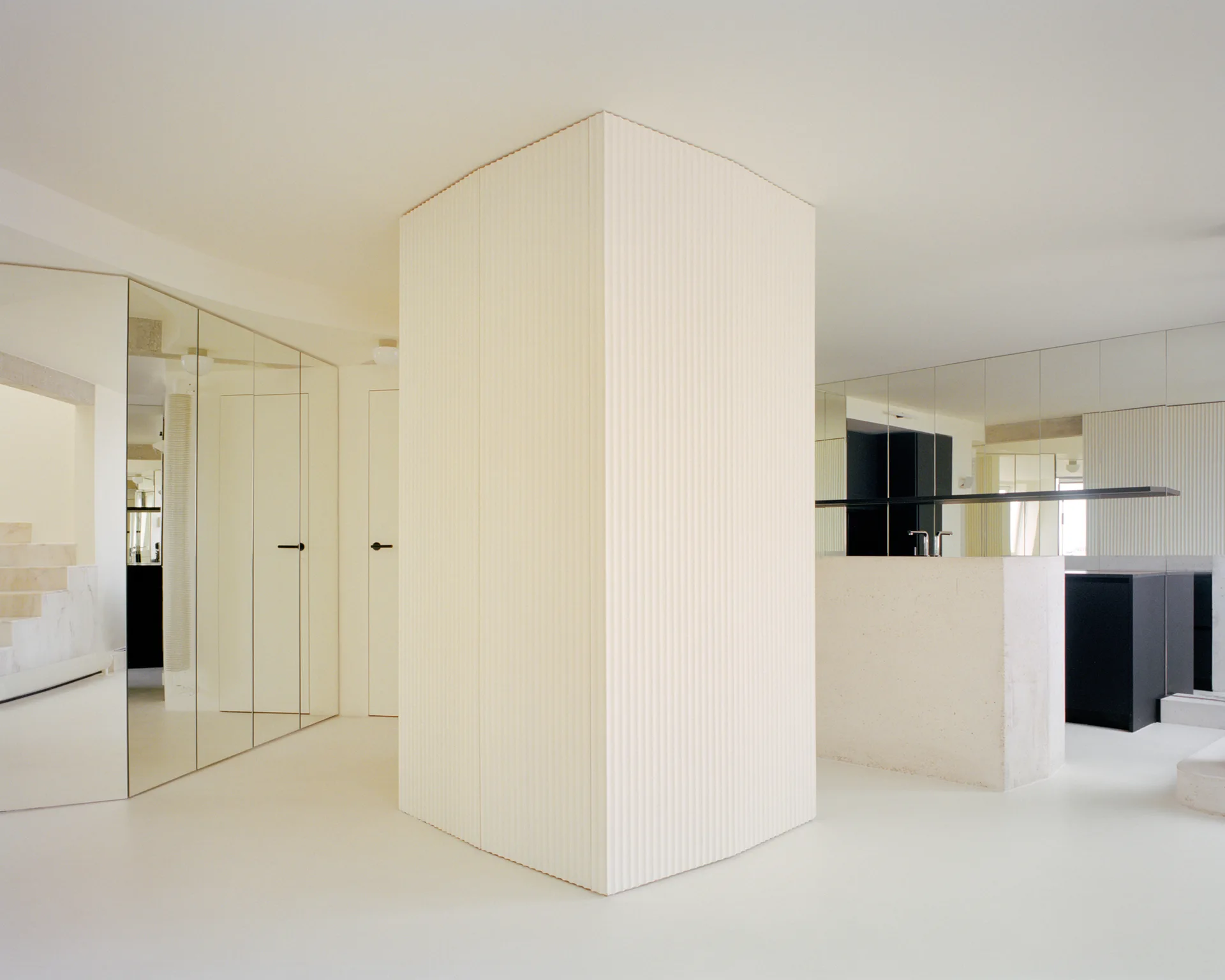 A minimalist white interior features a central fluted partition, mirrored wall panels, and a smooth stone-look island on a white floor.