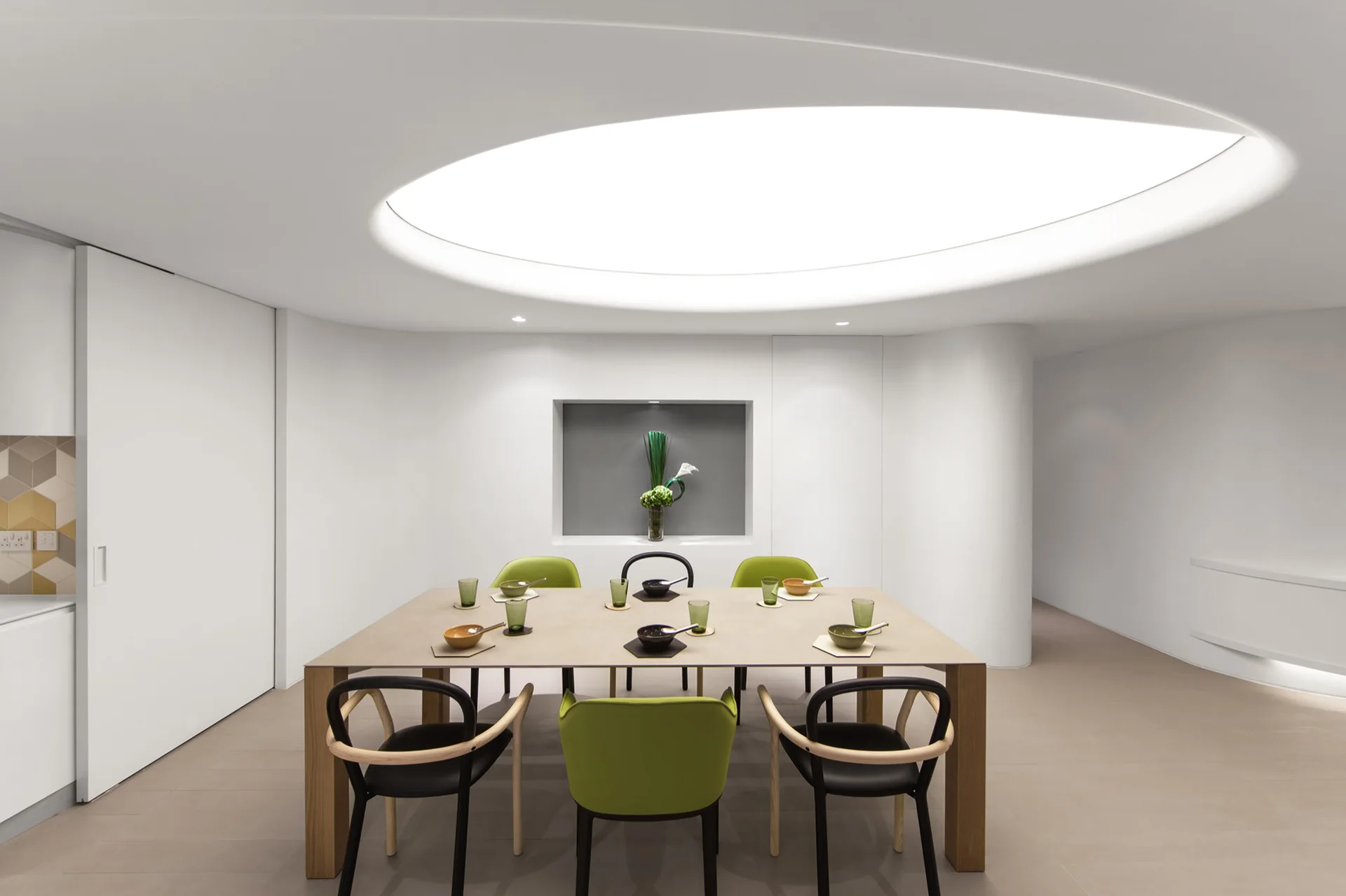 A modern dining room featuring a rectangular table with green and black chairs under a large, glowing circular ceiling light.