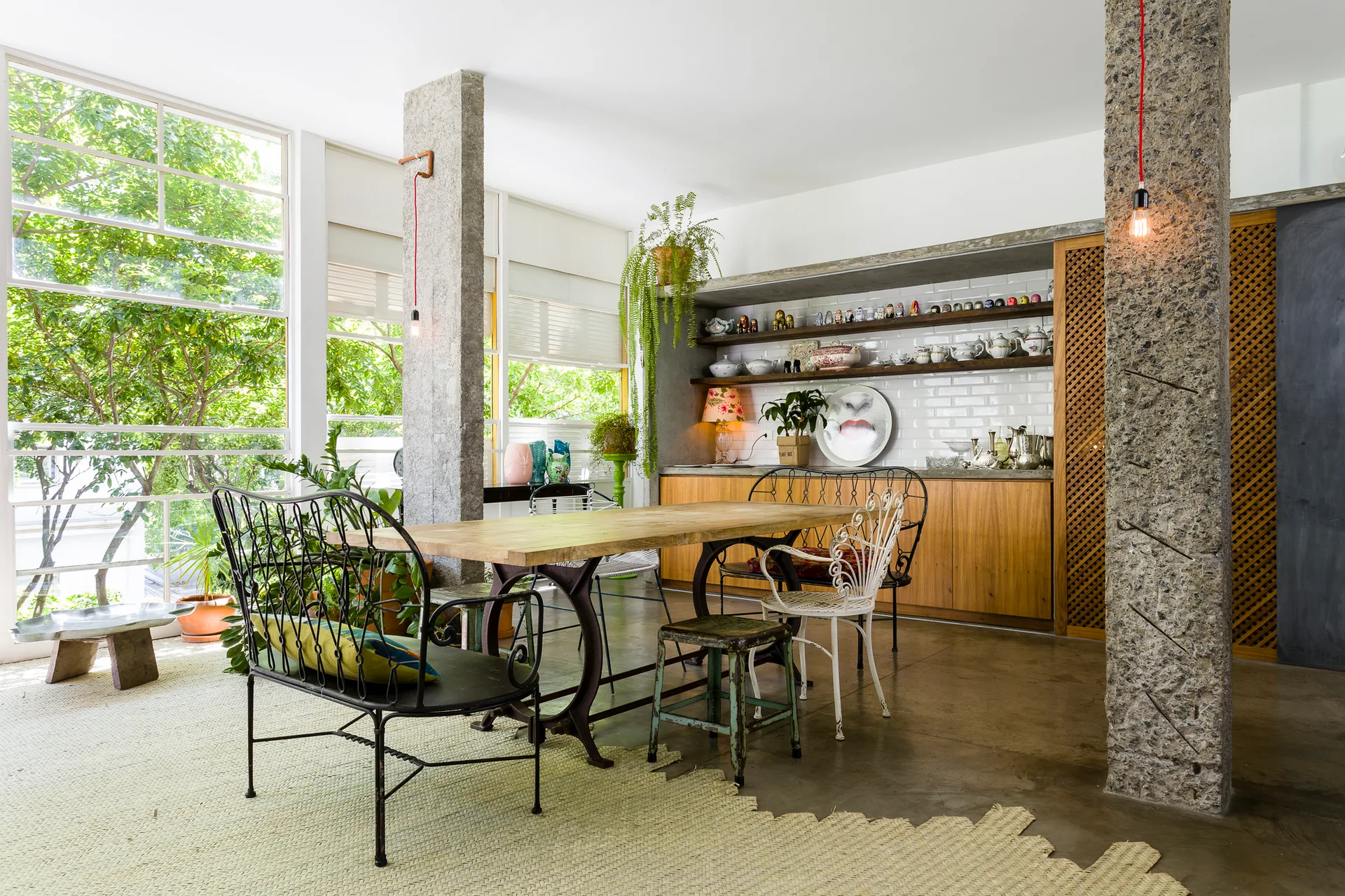 A long wooden dining table is surrounded by mismatched metal chairs and concrete pillars next to large windows overlooking green trees.