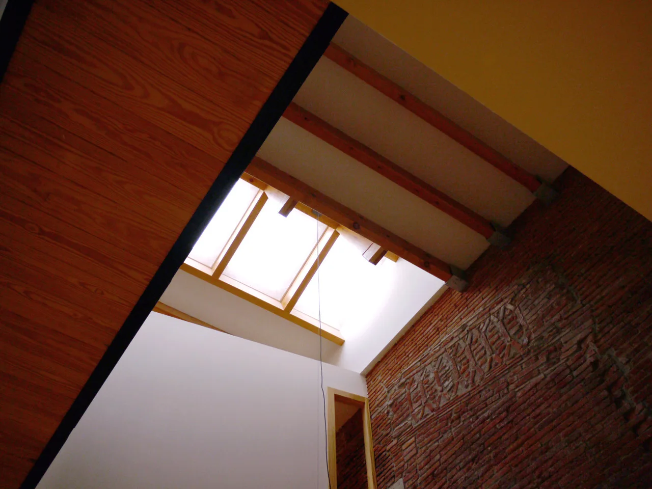 An upward view of a rectangular skylight in a white ceiling with exposed wooden beams alongside a textured red brick wall.