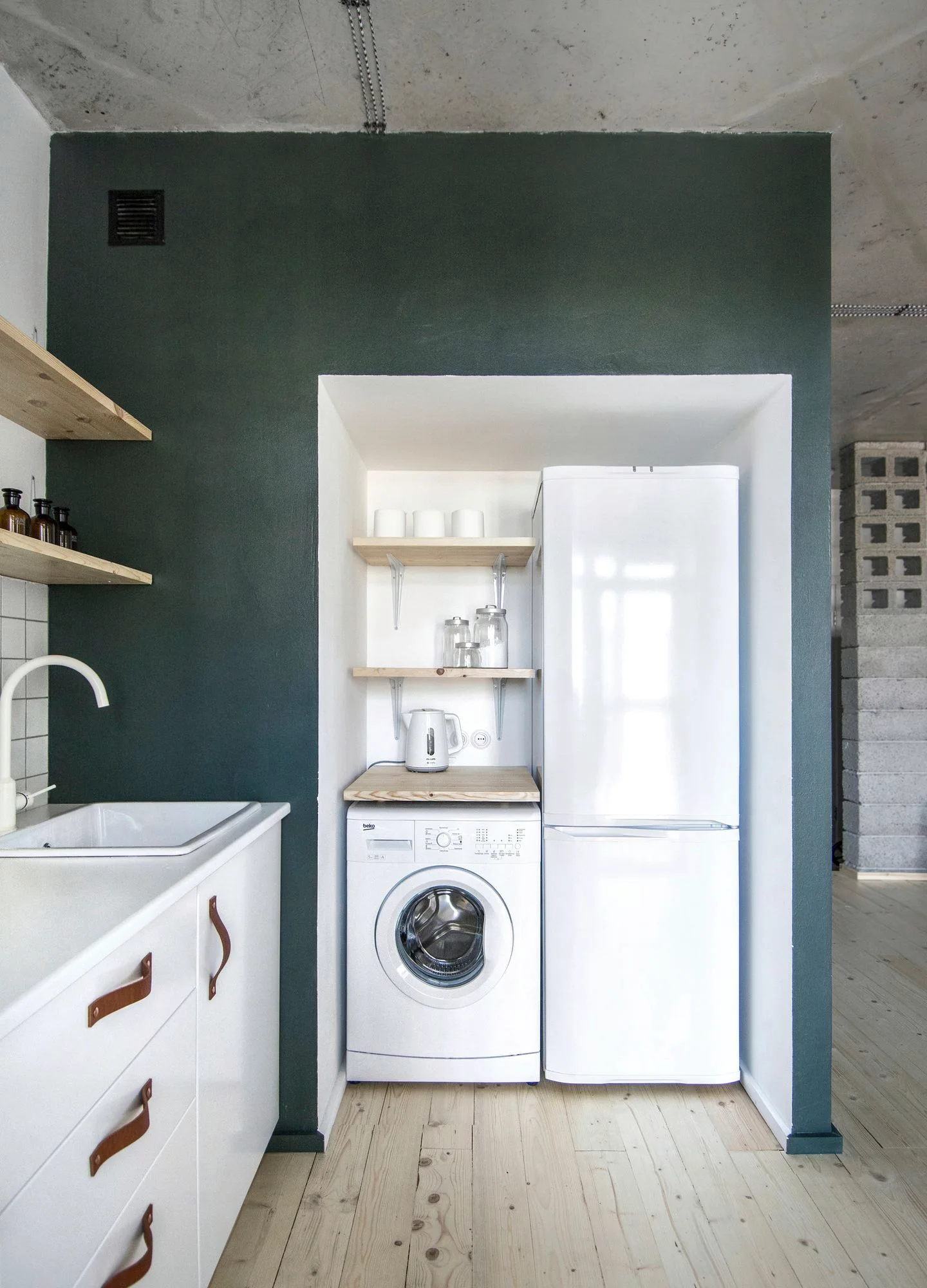 A compact kitchen features white appliances tucked into an alcove within a dark green wall next to white cabinets with leather pulls.