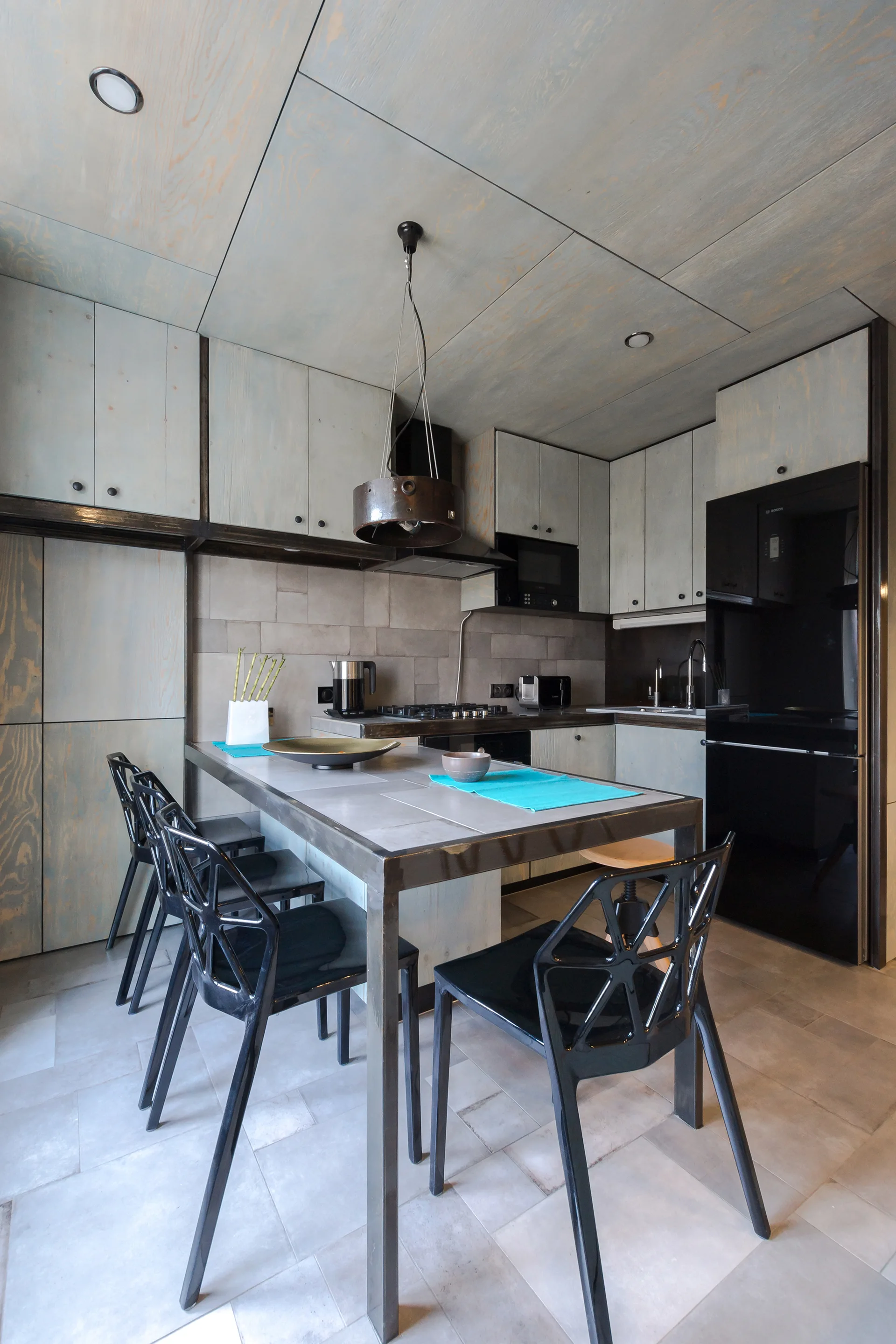 A modern kitchen with grey wood-paneled cabinets and ceiling, featuring a metal-framed dining island with geometric black chairs.