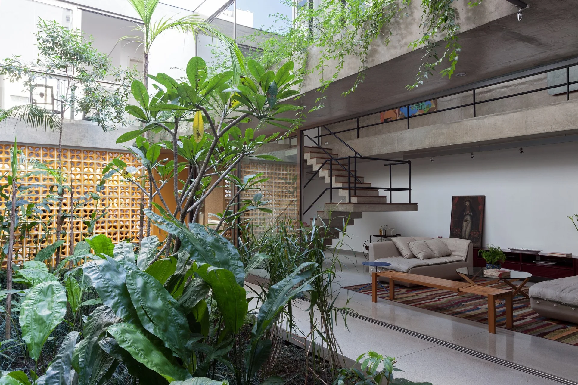 A modern living room features a floating concrete staircase, a beige sofa on a striped rug, and a lush indoor garden under a skylight.