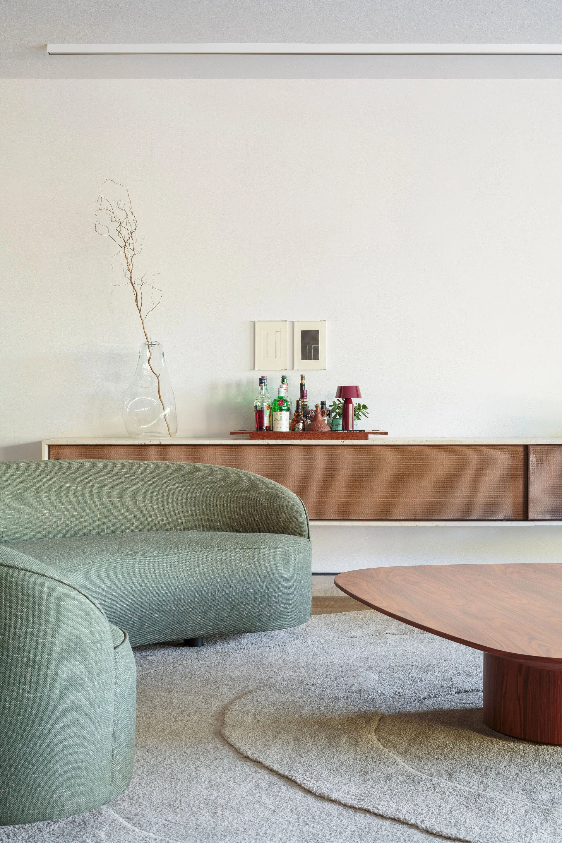 A living room featuring a curved sage green sofa and low wooden coffee table on a textured grey rug below a floating wooden sideboard.