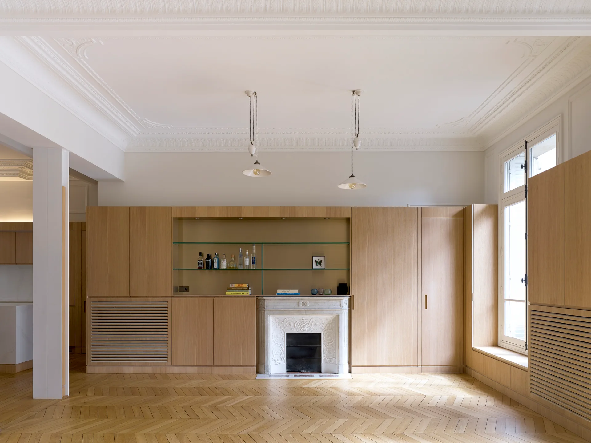 A spacious room features light oak built-in cabinetry integrated around an ornate white stone fireplace, set over herringbone hardwood floors.