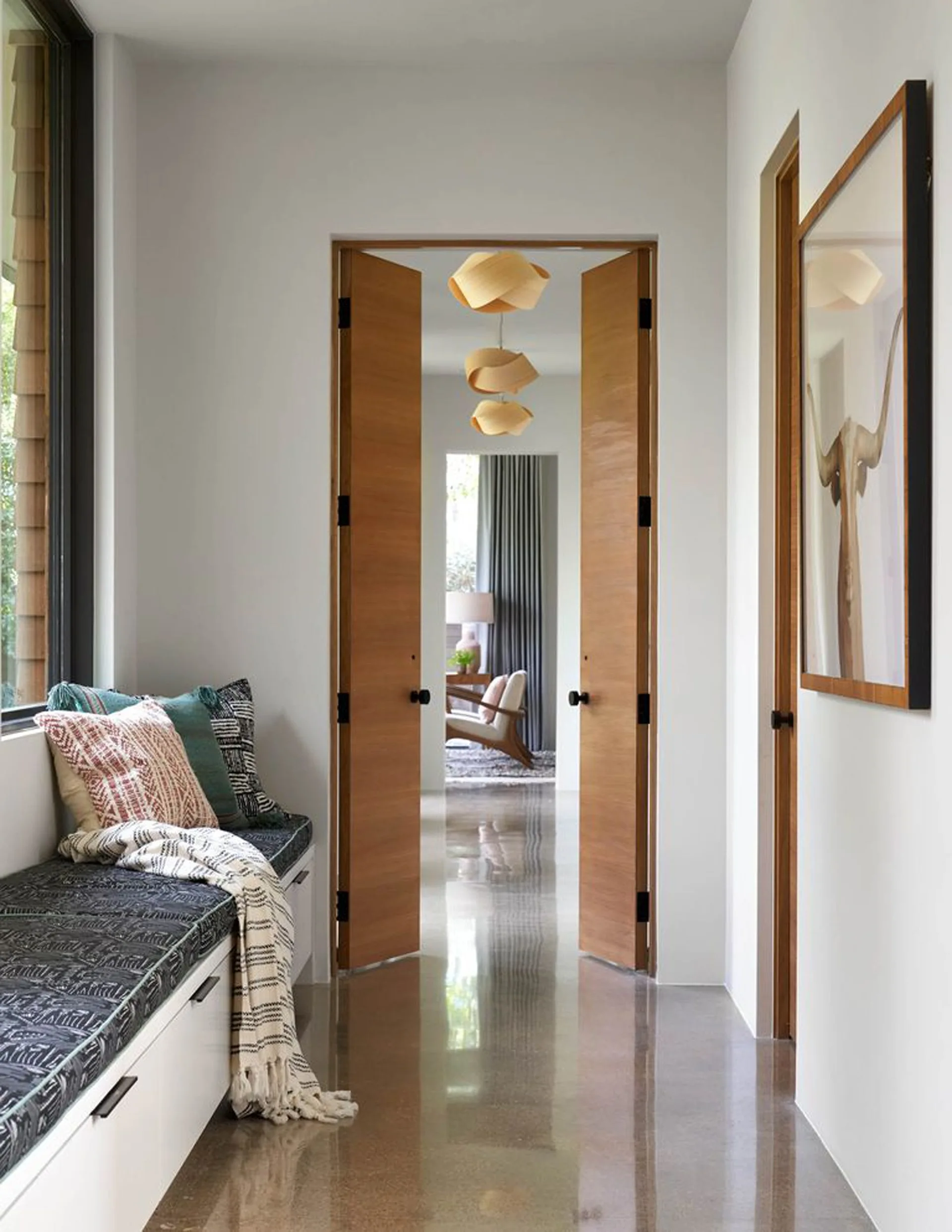 A bright hallway features a built-in window bench with patterned pillows, polished concrete floors, and open wooden double doors.