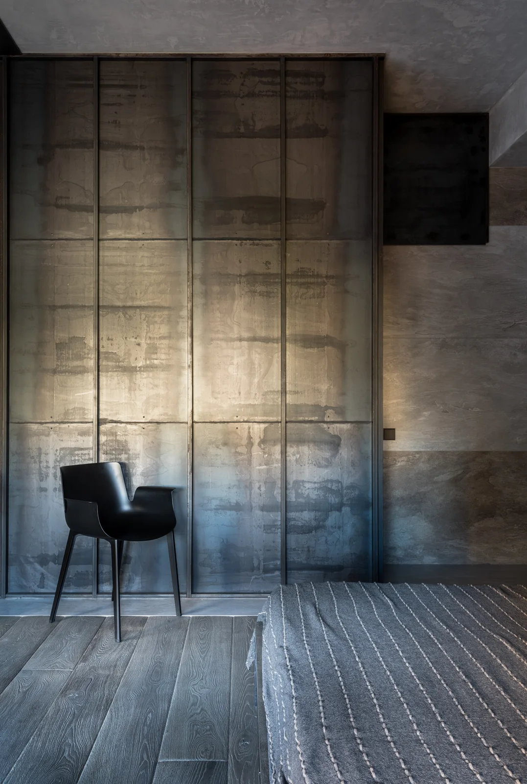 A minimalist bedroom featuring a textured gray metal partition wall, a black accent chair, and dark wood plank floors.