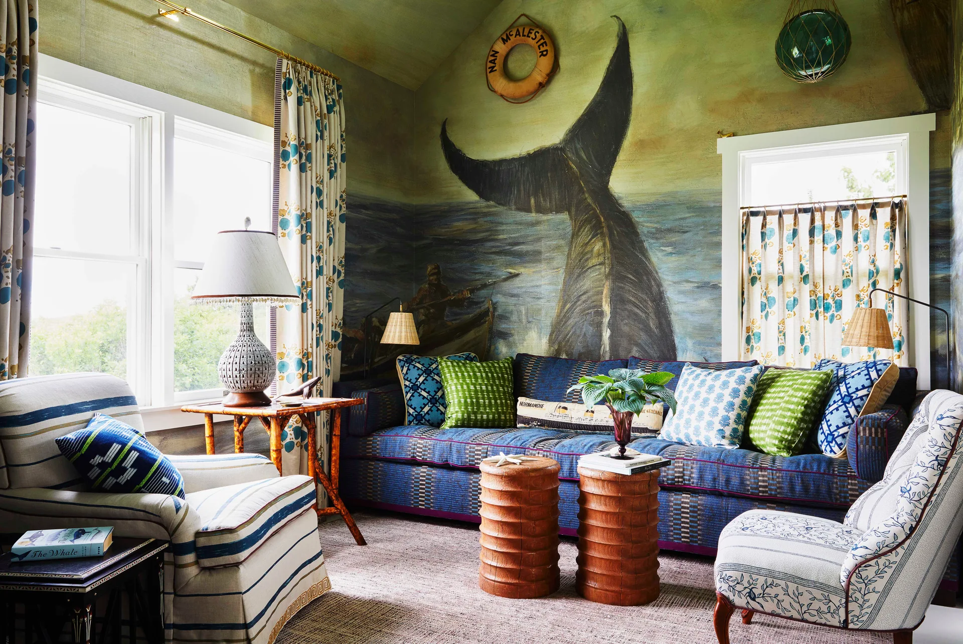 A coastal living room features a blue patterned sofa and striped armchair facing two wooden stool coffee tables in front of a painted whale mural.