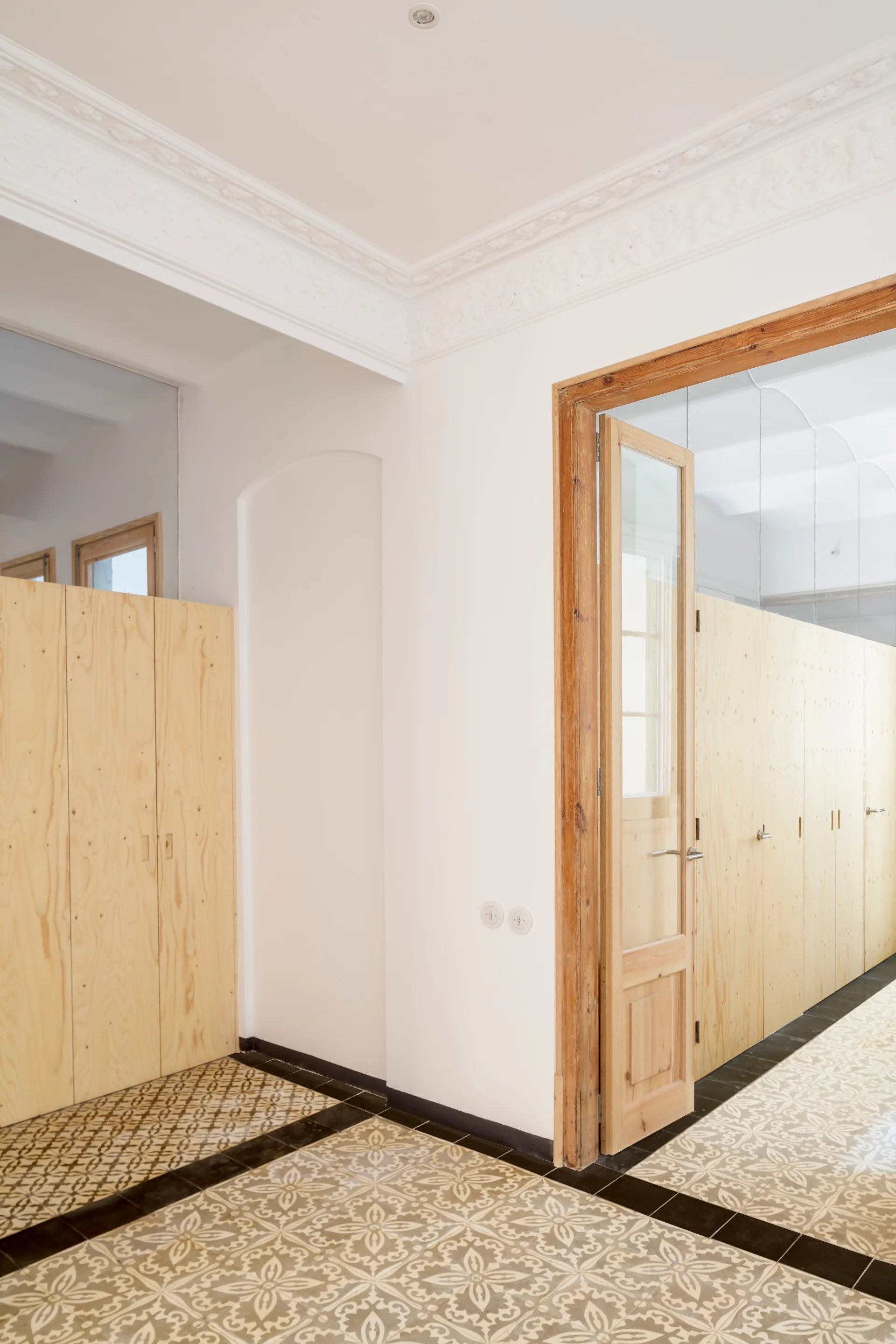 A bright hallway features light-toned plywood cabinets, patterned floor tiles, ornate white crown molding, and a natural wood glass-paneled door.