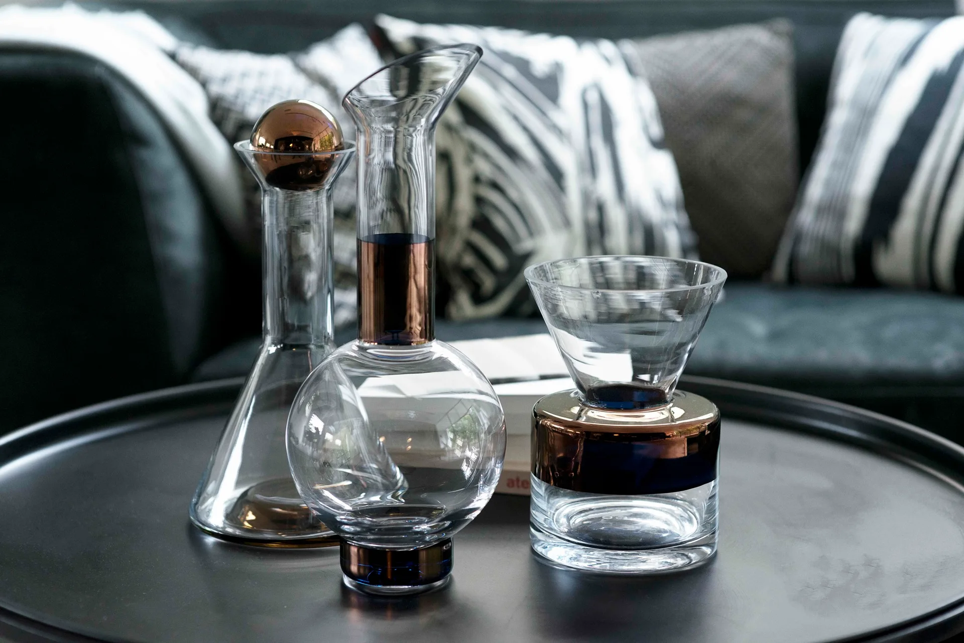 Three clear glass decanters with metallic bronze bands rest on a dark circular coffee table in front of a sofa.