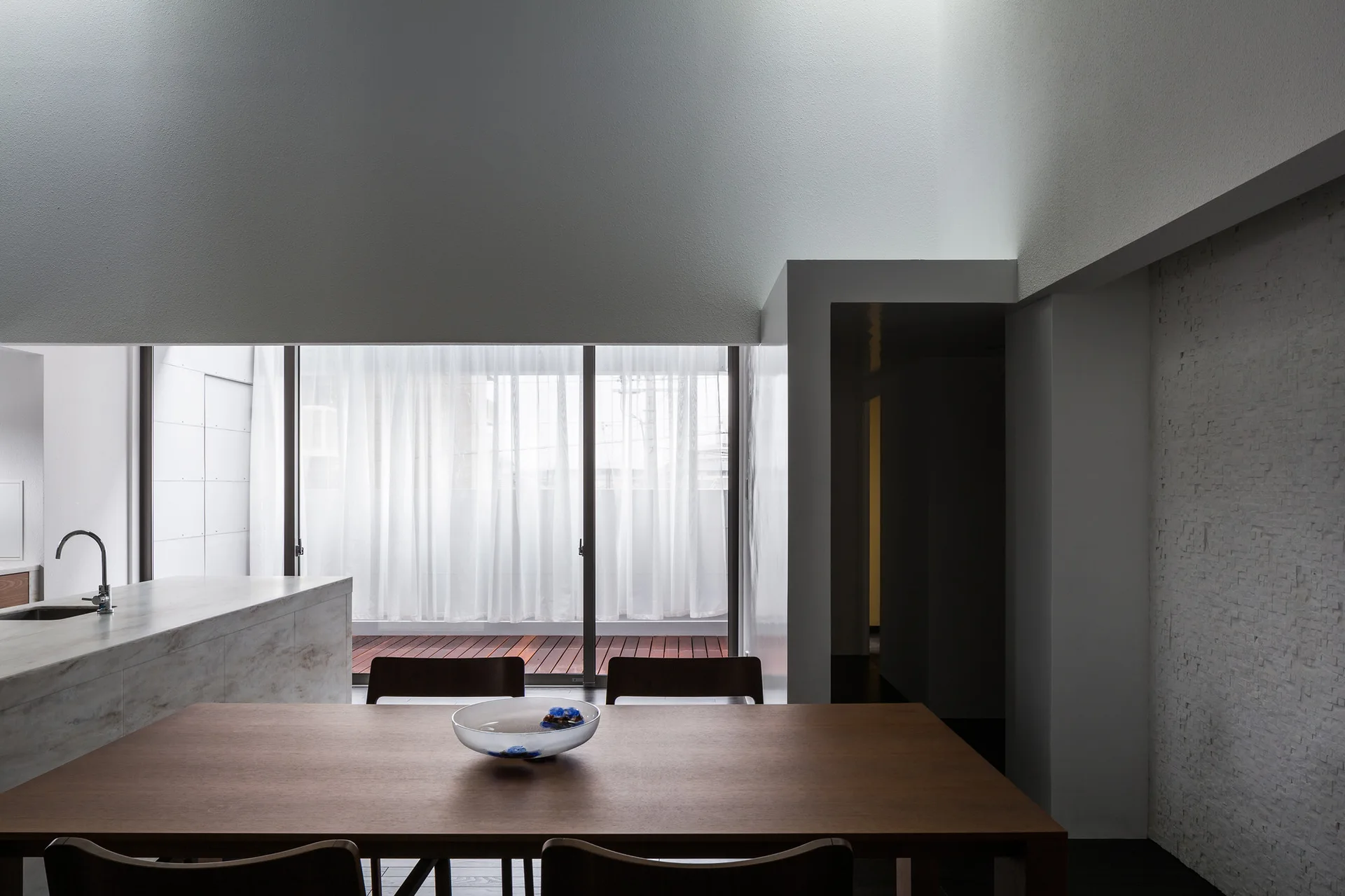 A minimalist dining room features a dark wood table and chairs next to a stone-patterned kitchen island and sliding glass doors opening to a deck.