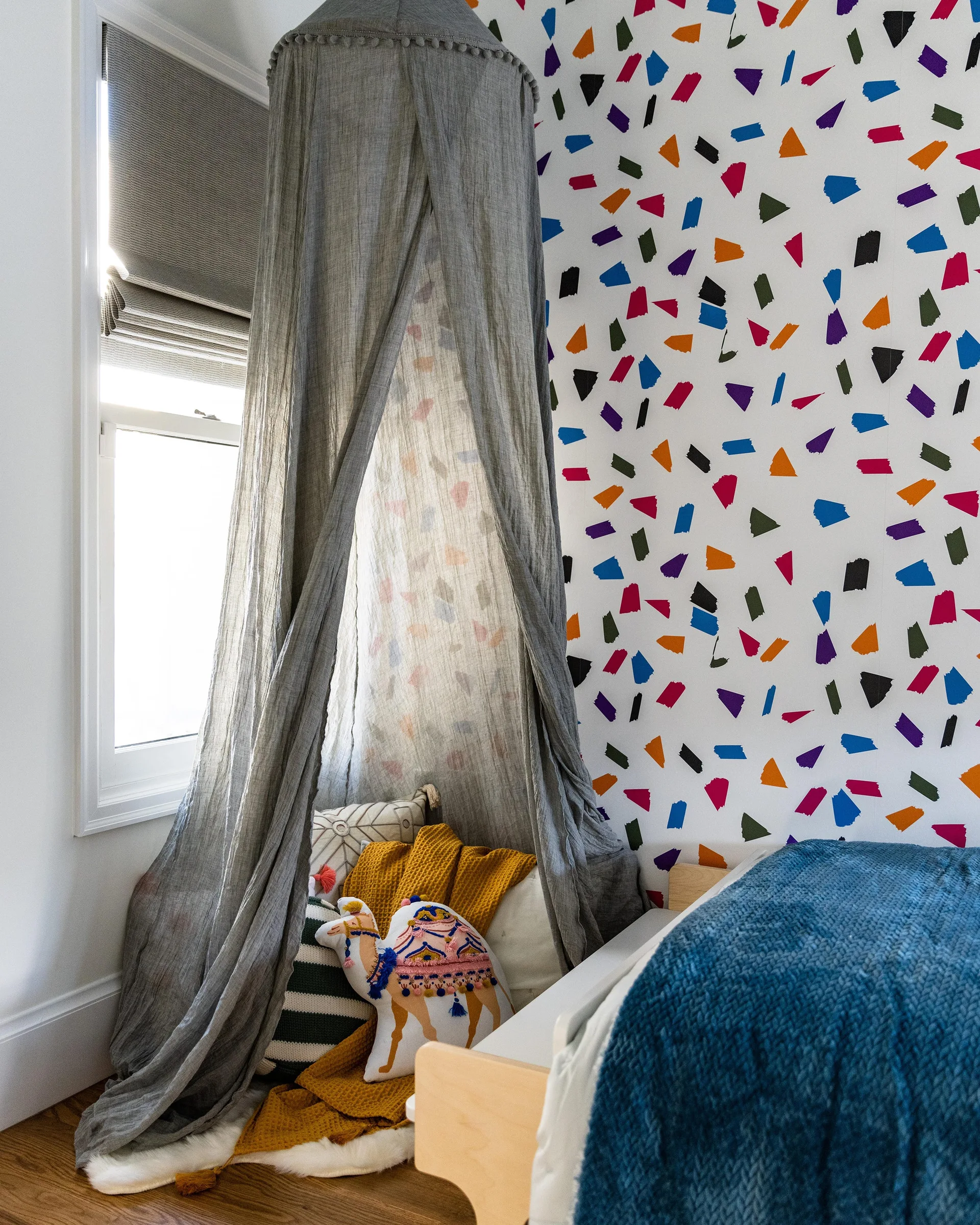 A child's bedroom corner with a grey fabric canopy over floor cushions, multi-colored brushstroke wallpaper, and a wooden bed frame.