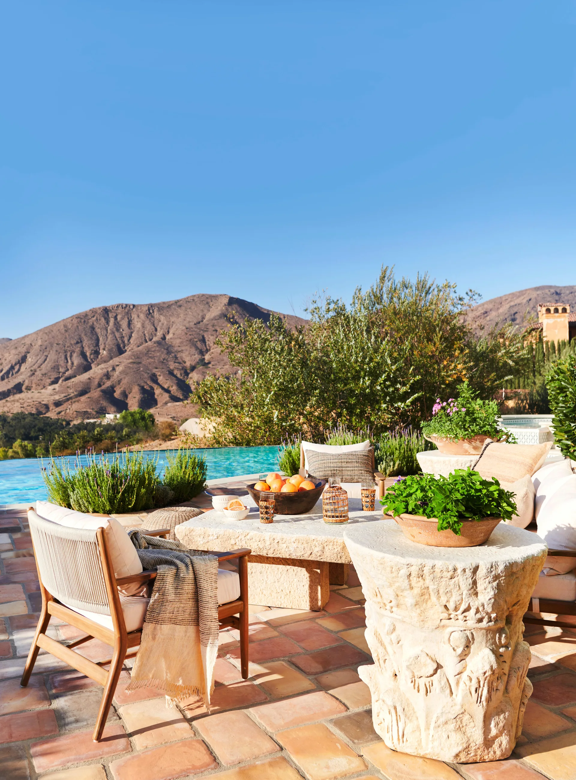 An outdoor patio featuring terracotta floor tiles, carved stone tables, wooden lounge chairs, and an infinity pool overlooking desert mountains.