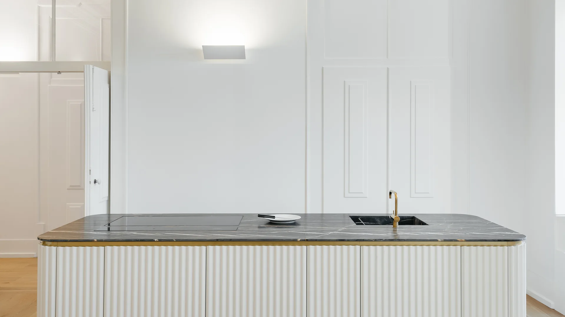 A minimalist kitchen island features a fluted white base, a dark veined stone countertop with an integrated sink, and a brass faucet.