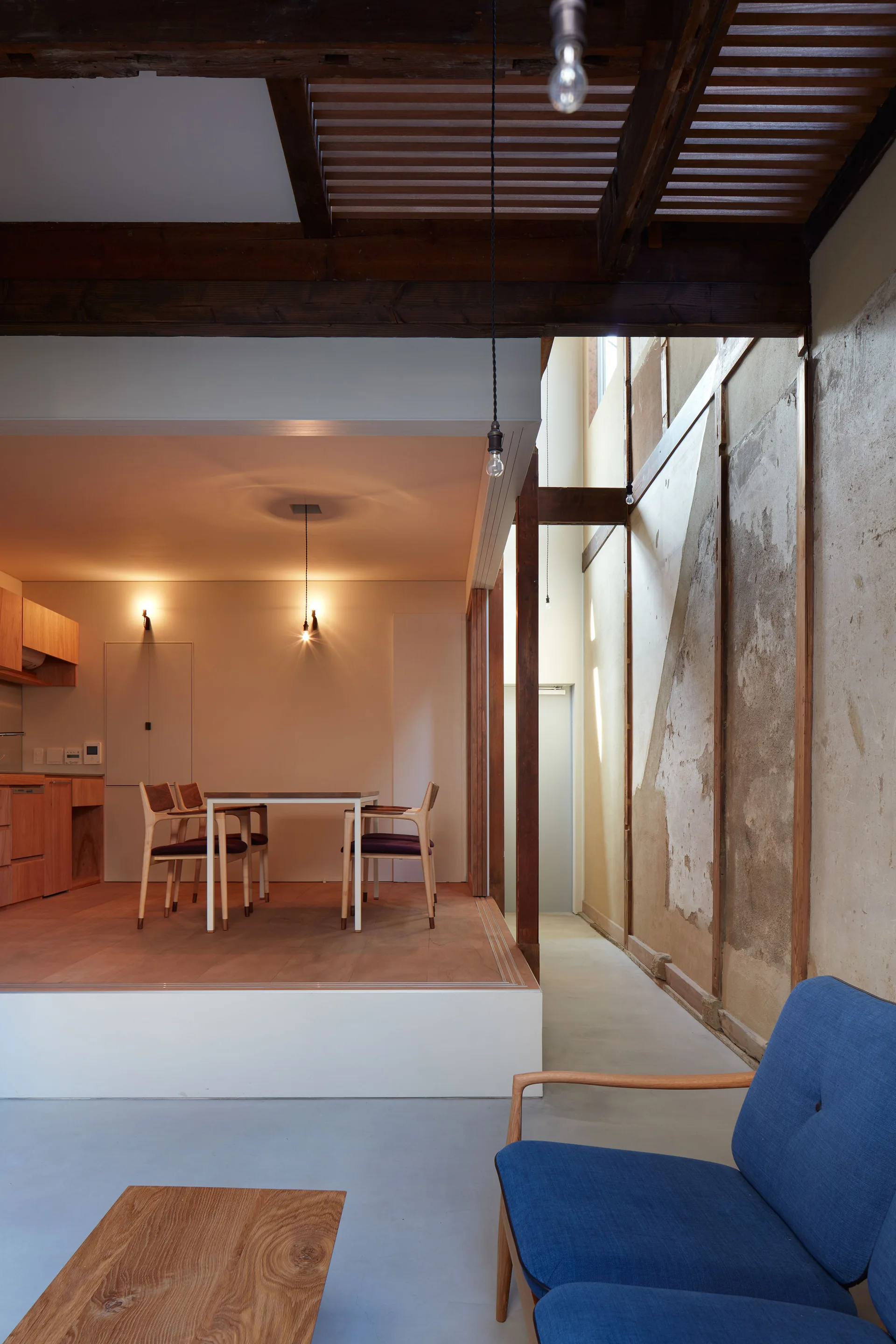 A split-level home features a raised wooden dining area next to a rough plaster wall, with a blue armchair in the concrete-floored foreground.