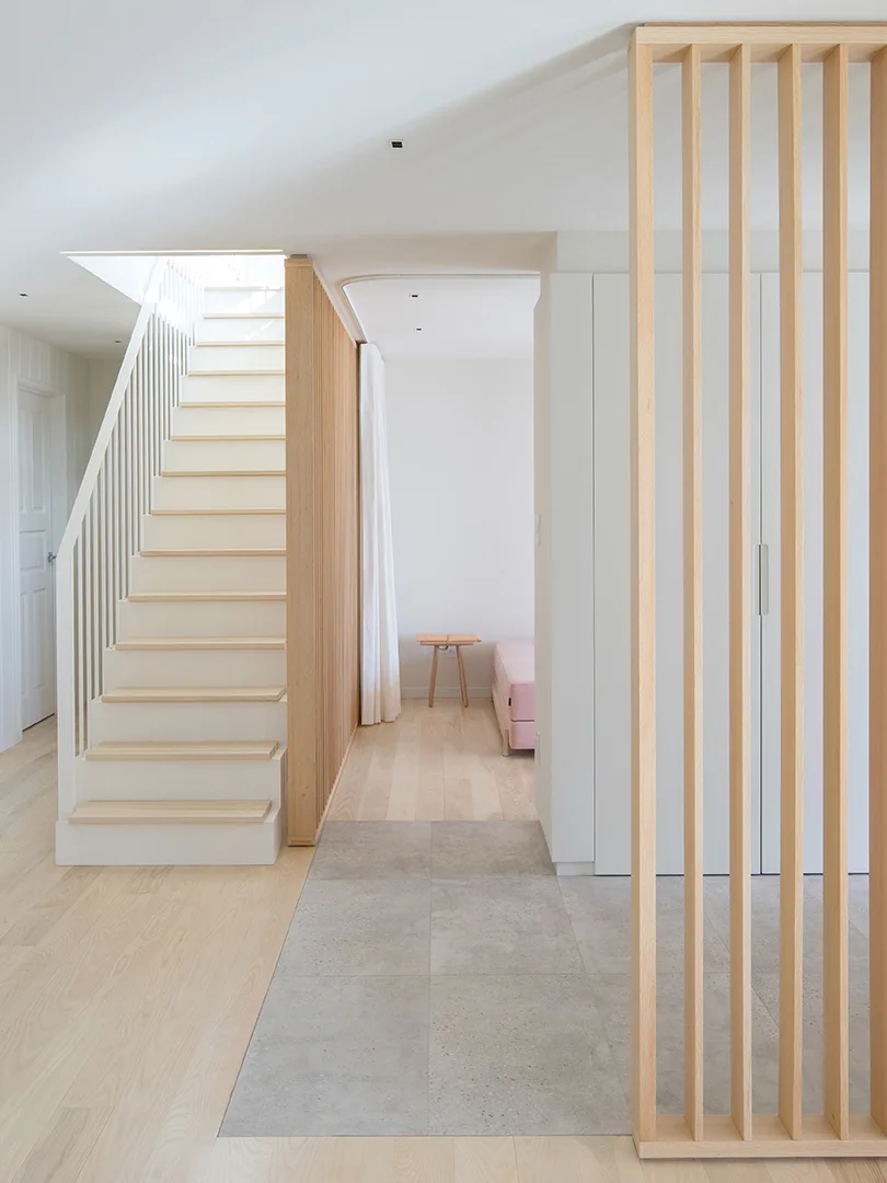 A minimalist entryway featuring a white staircase with wood treads, vertical wood slat screens, and a transition from gray tile to wood flooring.