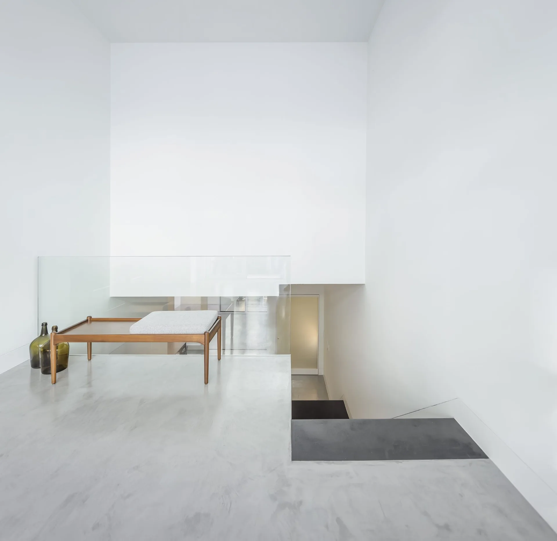 A minimalist staircase landing with light grey concrete floors features a mid-century wooden bench and a clear glass railing against white walls.