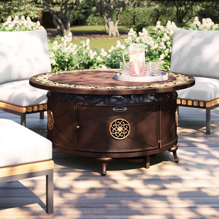 A circular bronze fire pit table featuring gold scrollwork details sits on a wooden deck surrounded by white cushioned outdoor armchairs.