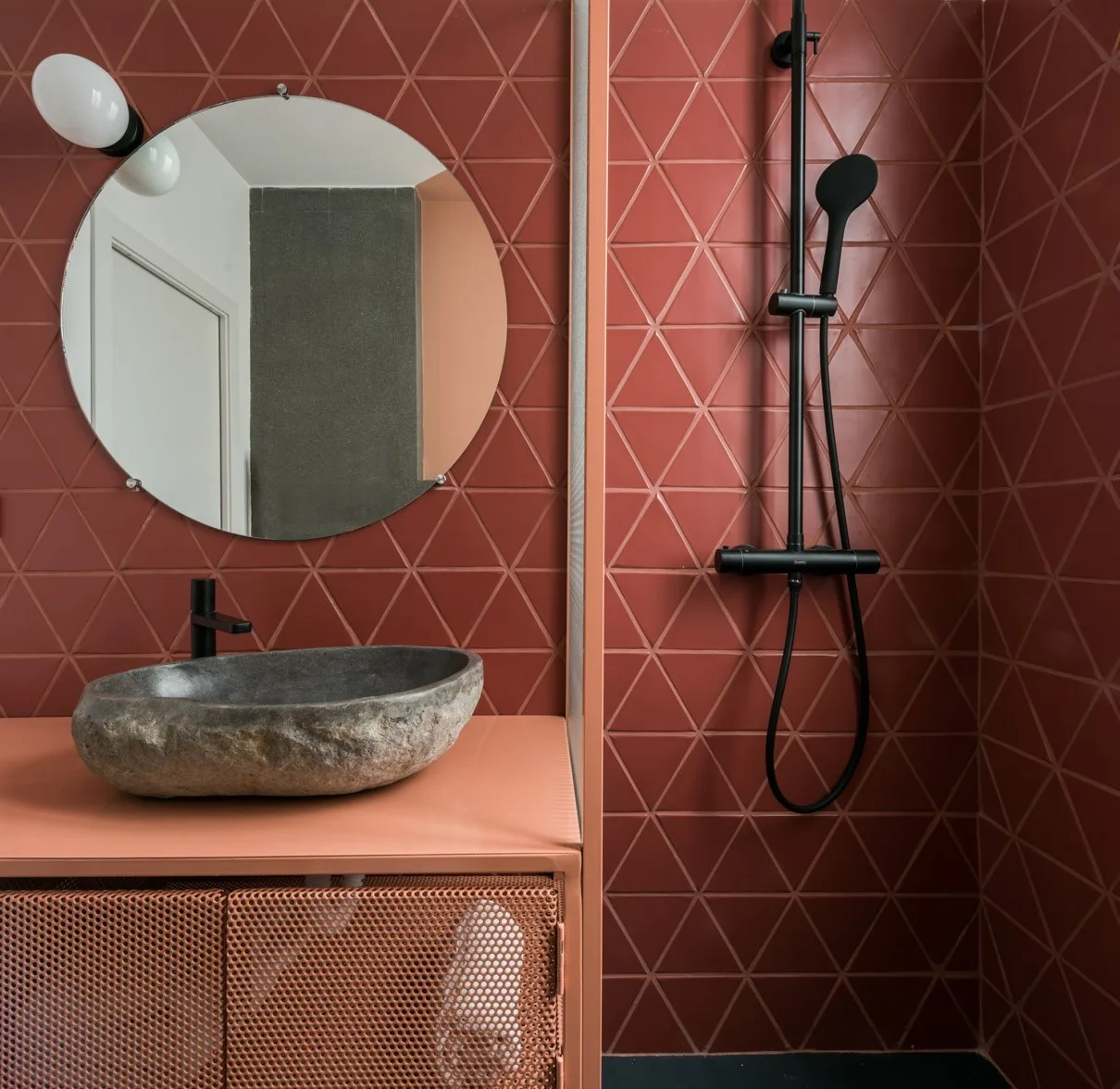 A modern bathroom features triangular terracotta wall tiles, a rough stone vessel sink, a round mirror, and matte black fixtures.