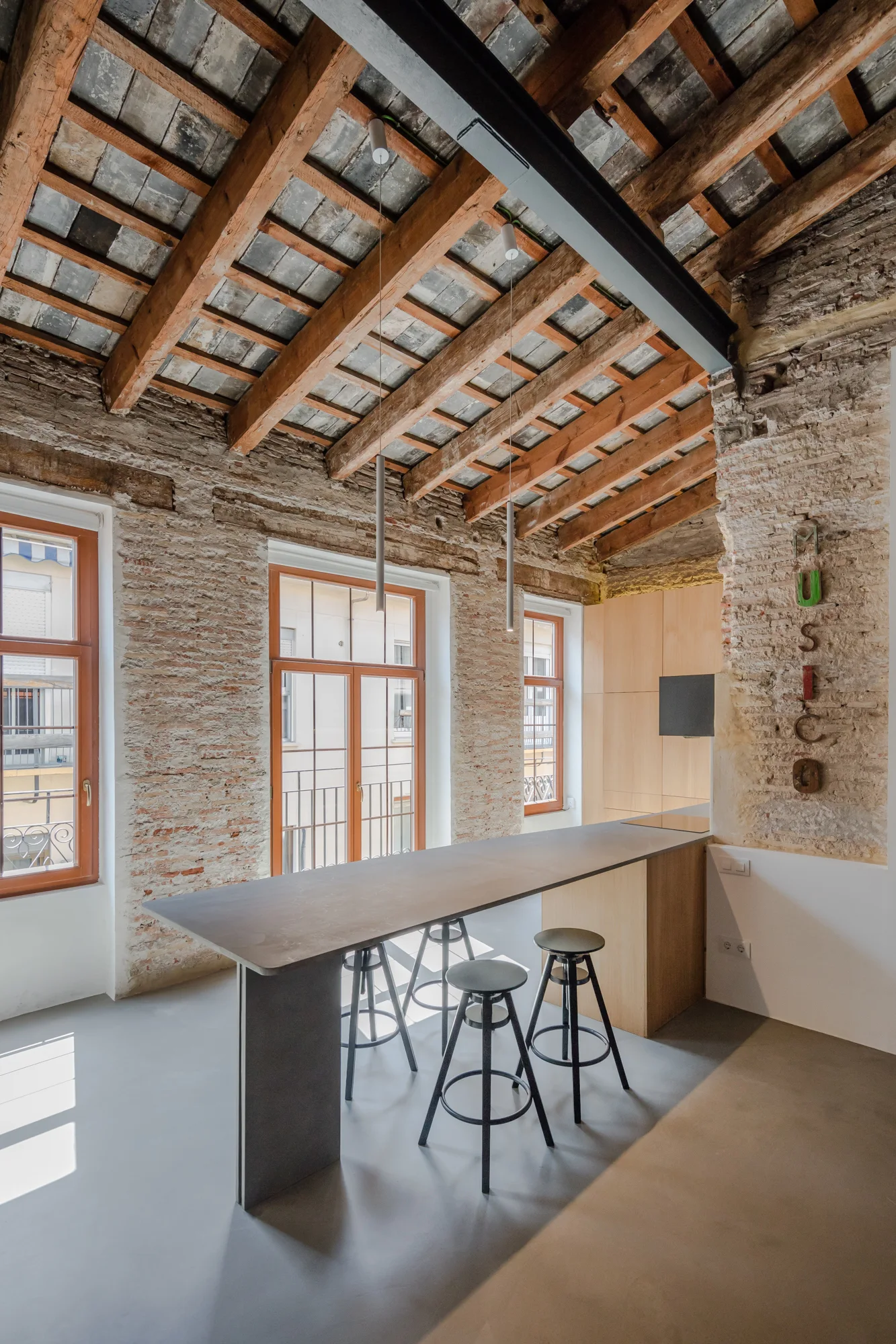 A modern gray kitchen island with black stools stands in a room featuring rugged exposed brick walls and an original wooden beam ceiling.