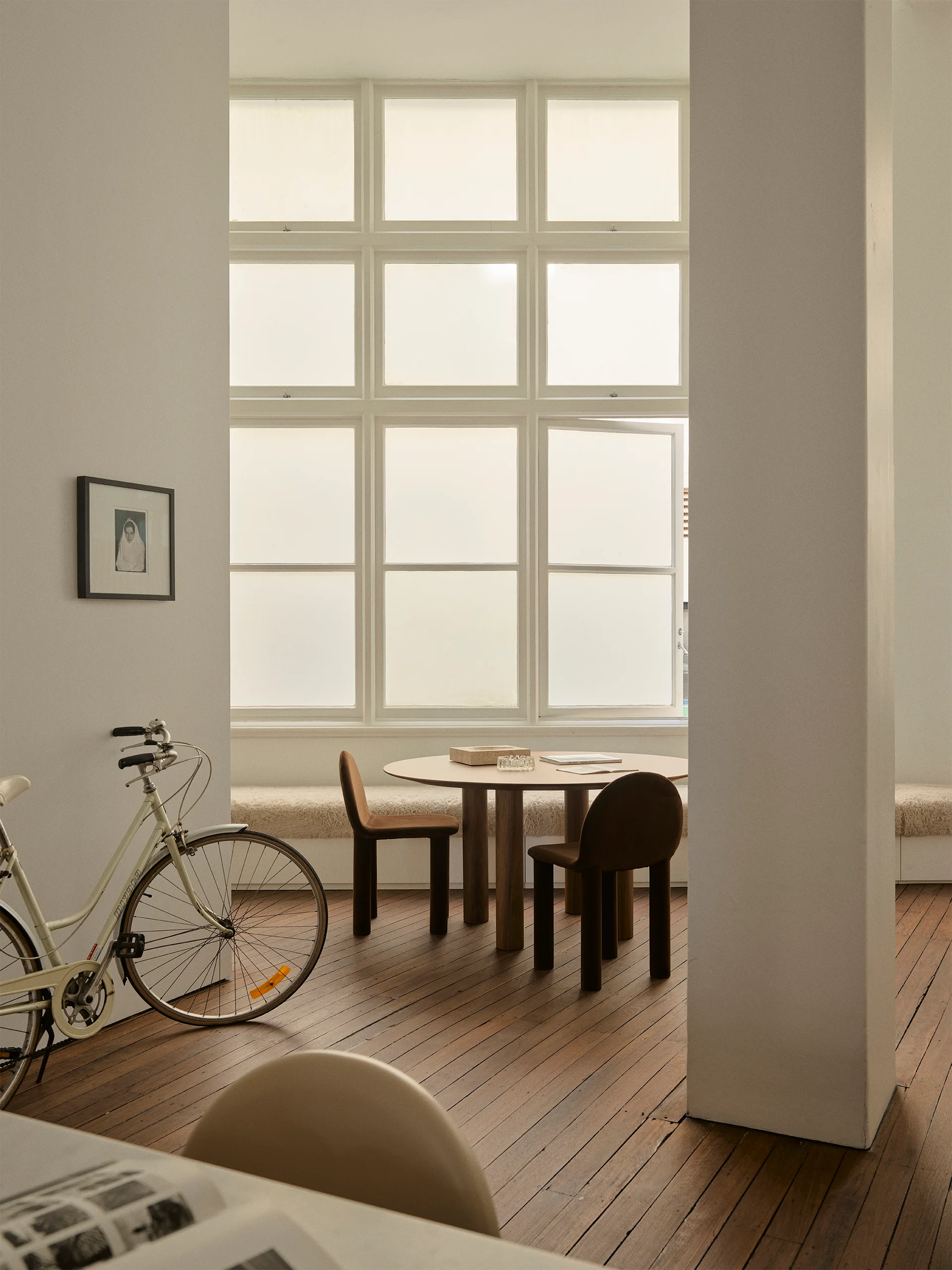 A round wooden dining table with two brown chairs sits next to a long built-in window bench beneath a large multi-pane grid window.