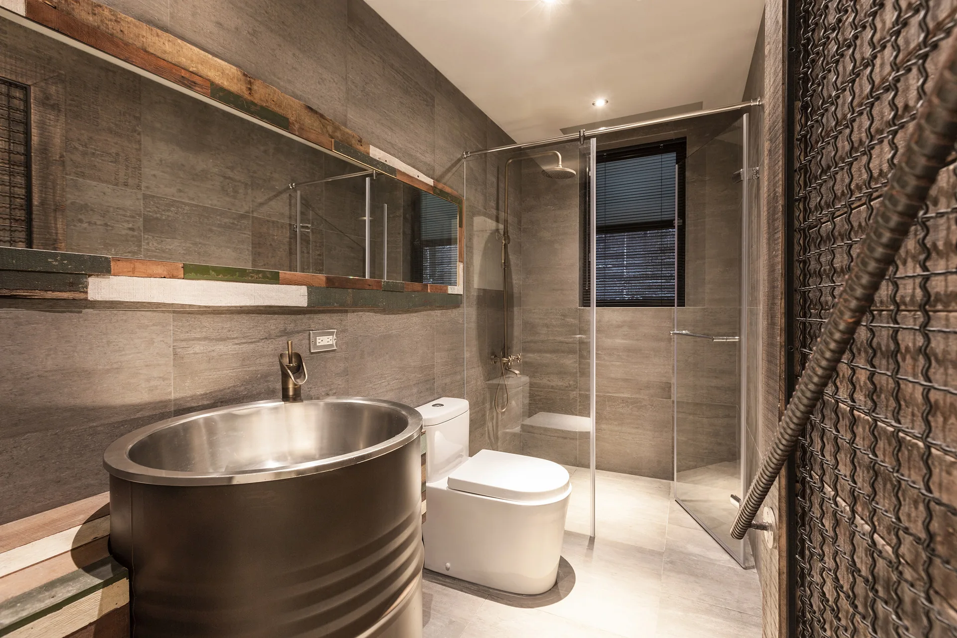 An industrial bathroom features a metallic barrel sink, a modern toilet, large grey wall tiles, and a glass-enclosed walk-in shower.