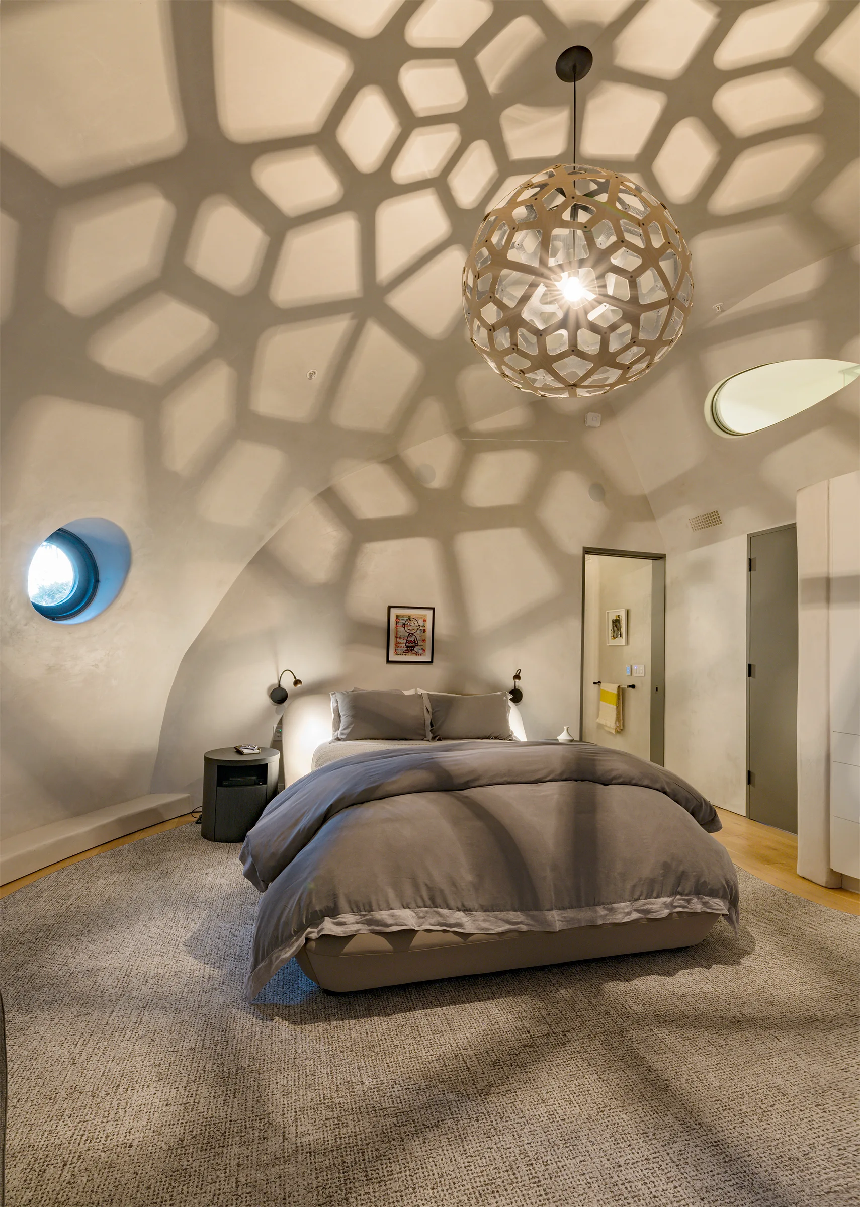 A minimalist bedroom beneath a plaster dome ceiling features a bed with grey linens and a spherical pendant lamp casting geometric shadows.