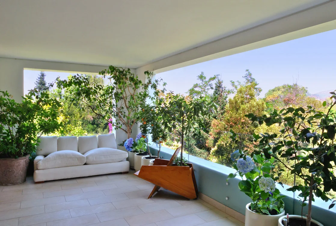 A covered terrace features a white outdoor sofa, an angular wood planter, and numerous potted plants along a glass railing.